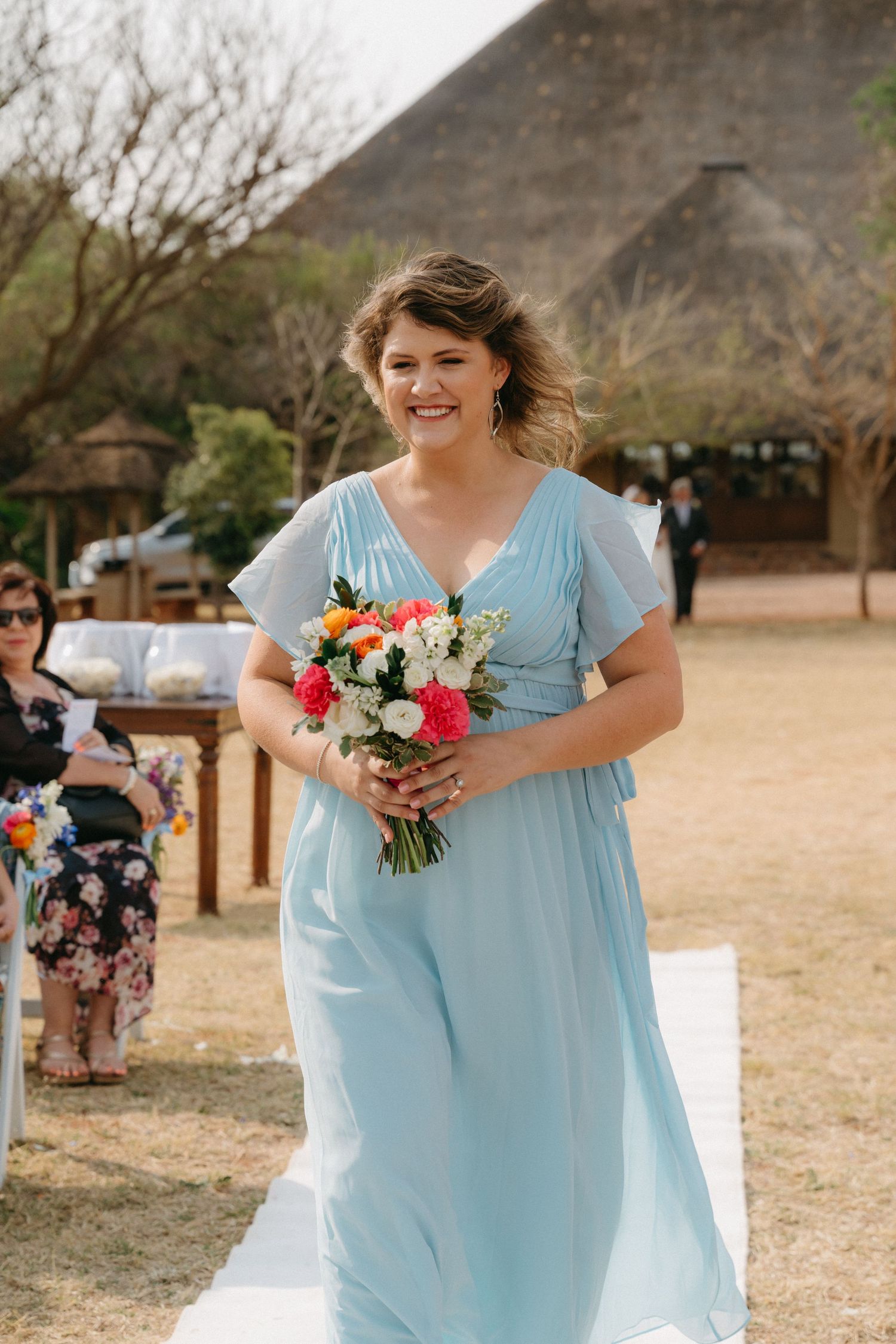 Bridesmaids in powder blue dresses carry vibrant bouquets during an outdoor wedding ceremony.