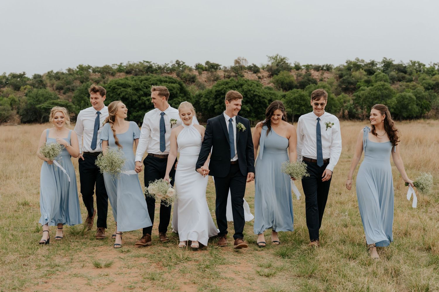 Wedding party in light blue bridesmaid dresses and dark suits posed together in a sunny outdoor field.