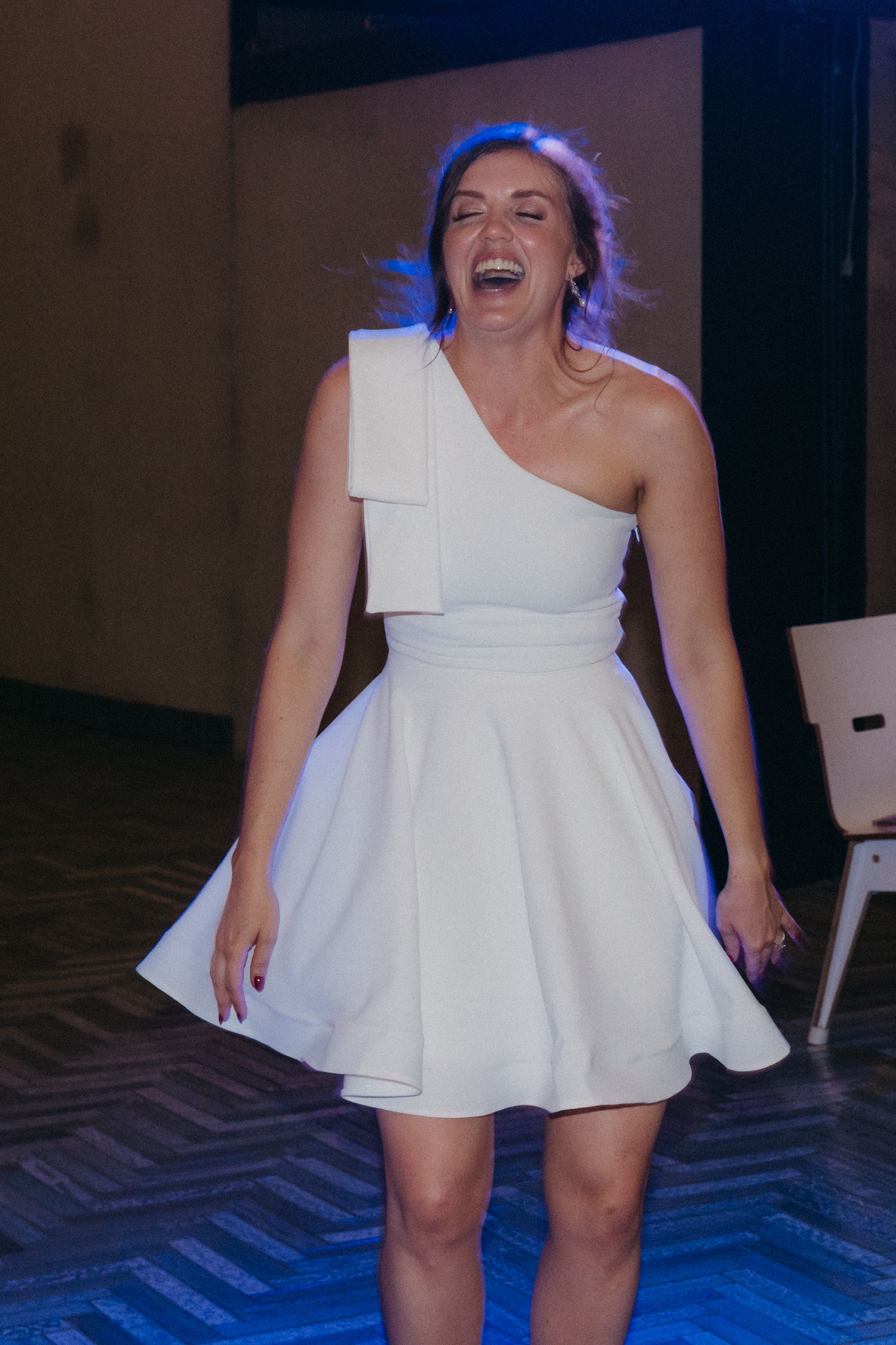 A person in a one-shoulder white dress laughs joyfully on a dance floor with blue lighting.