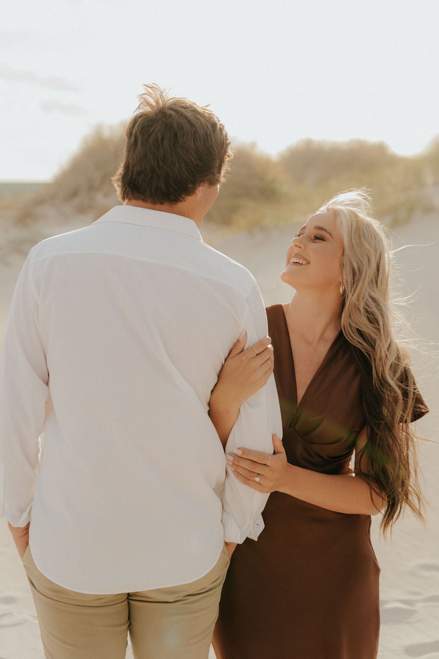 A couple shares a joyful moment on a sandy beach, woman in brown dress laughing as man in white shirt turns toward her.