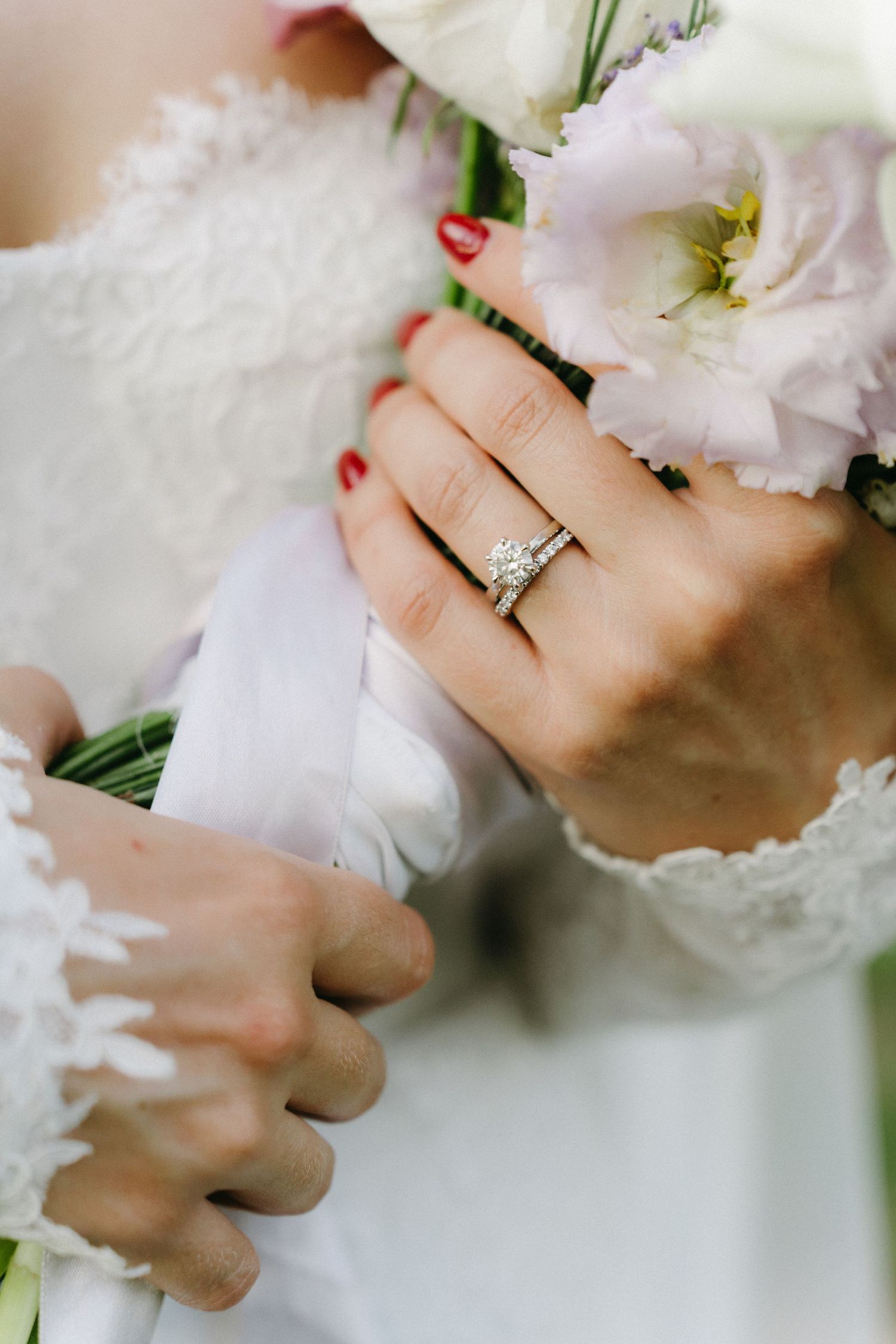 A close-up shot of hands holding white flowers with a sparkly diamond engagement ring and red nail polish.