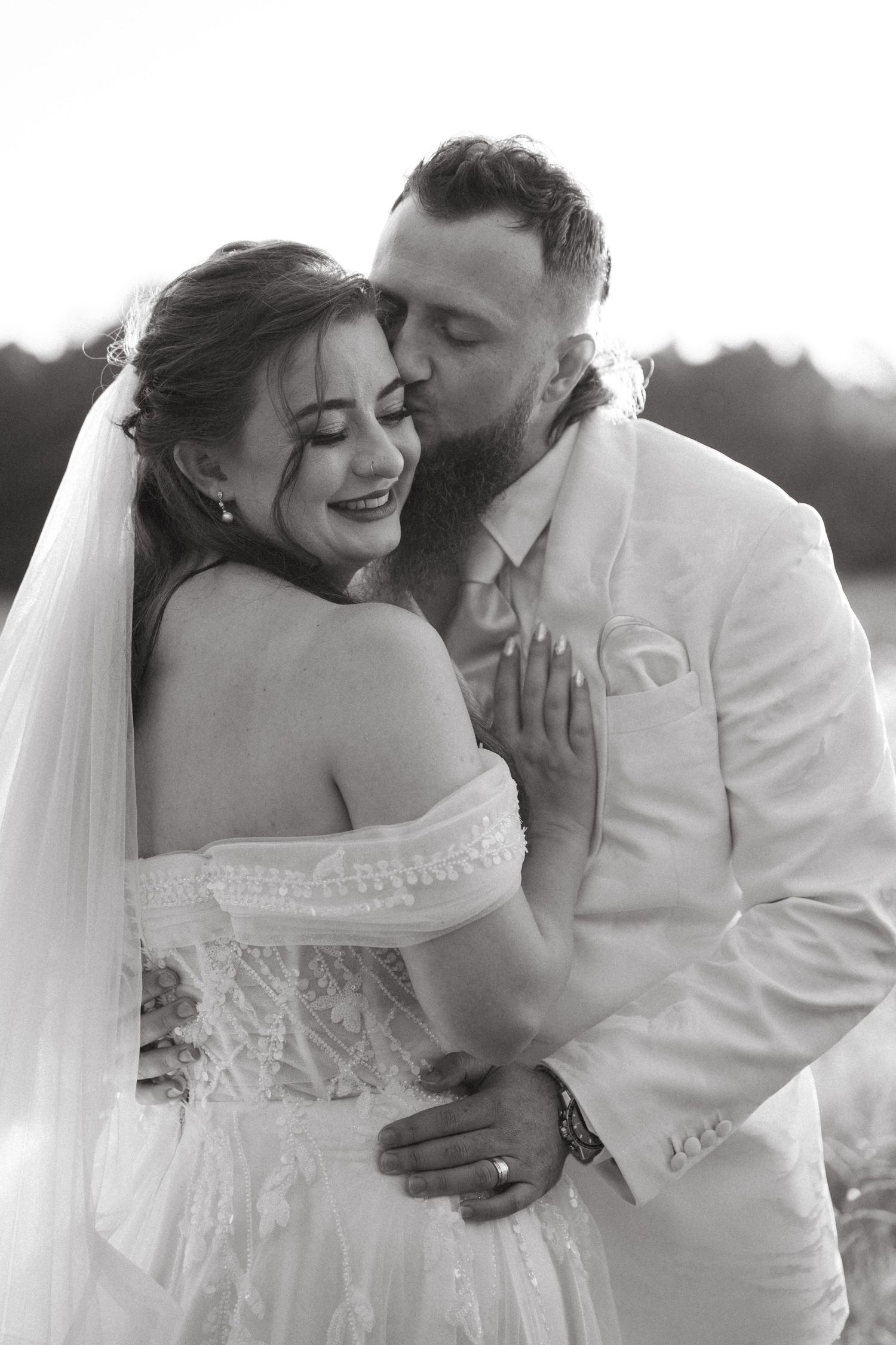 A romantic black and white photo of newlyweds embracing outdoors, with the bride in a strapless lace gown.