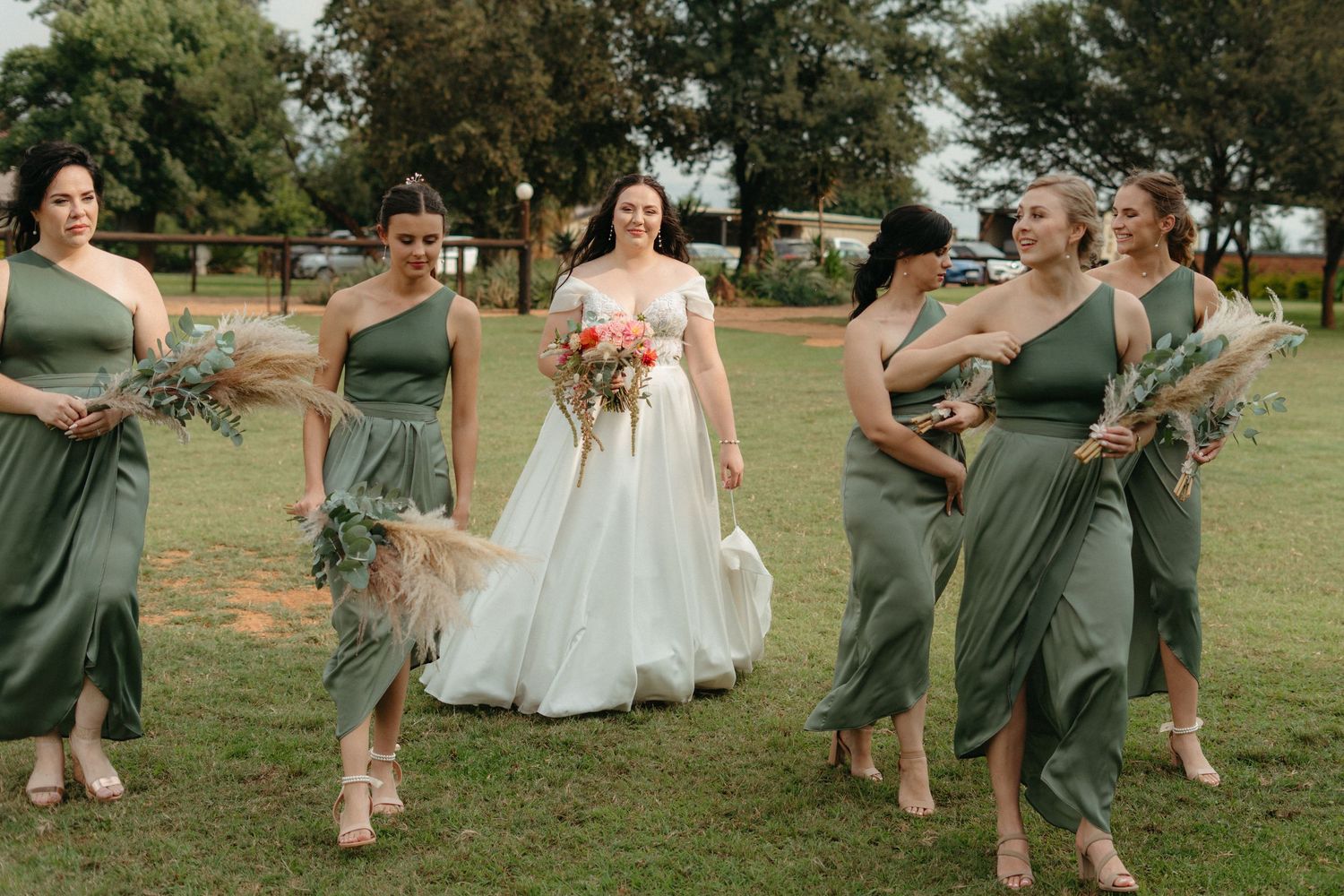 Bridesmaids in sage green dresses dance and laugh together on a grassy lawn at an outdoor wedding celebration.