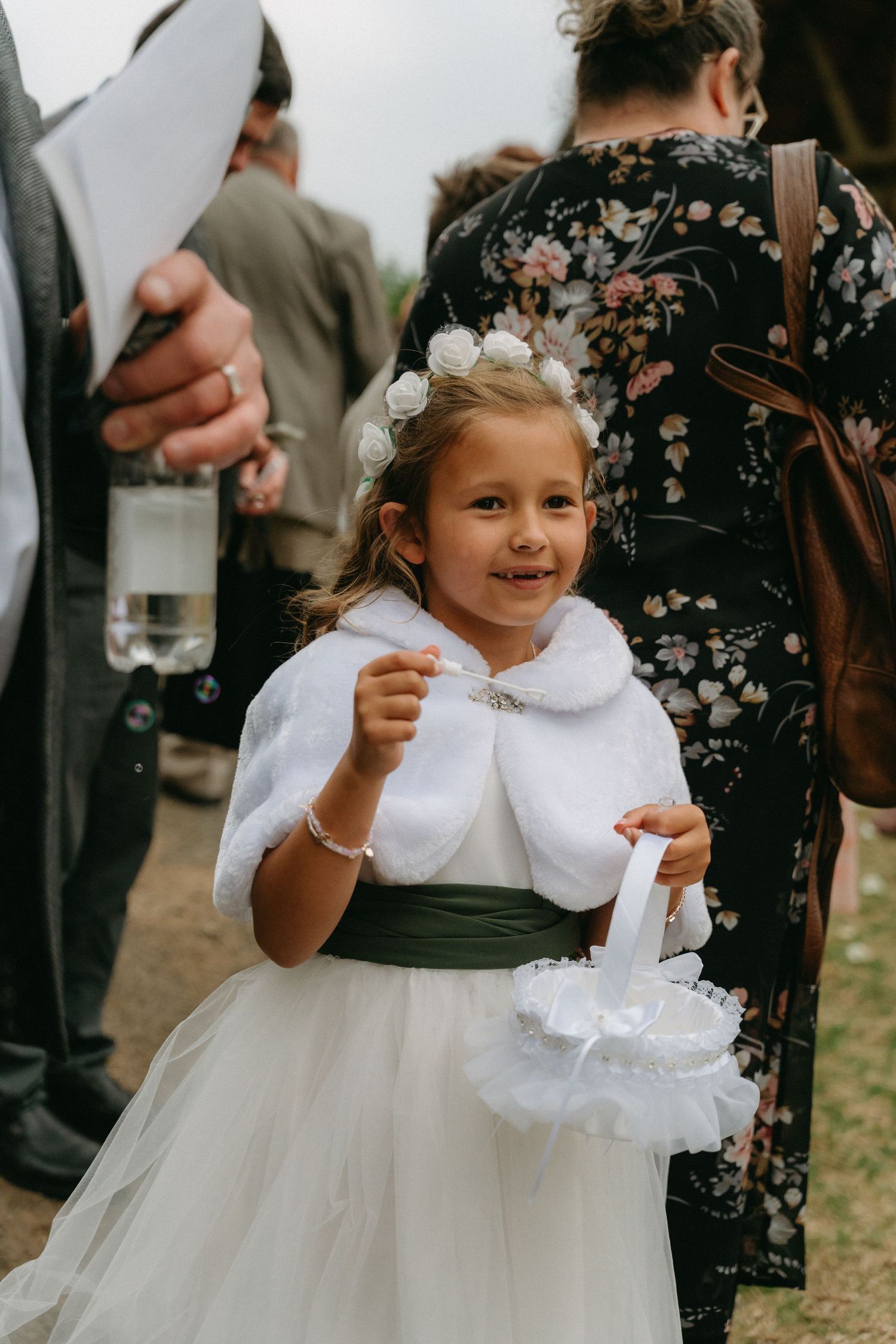 Young flower girl in white dress and black sash smiles at outdoor wedding.