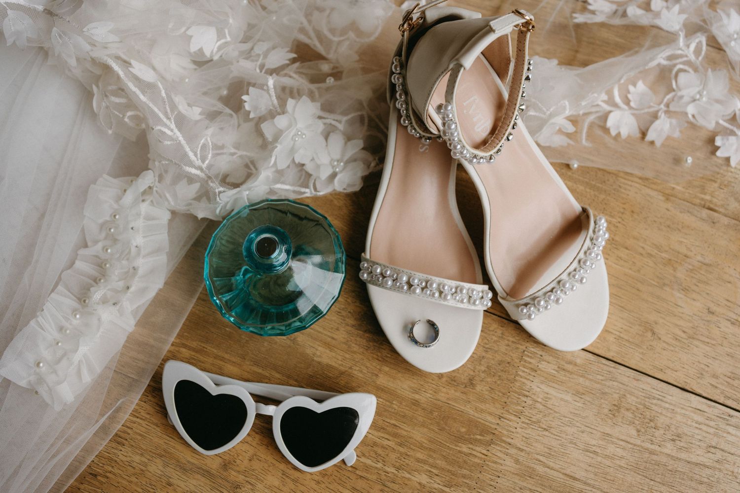 Wedding accessories including jeweled flat sandals, heart-shaped sunglasses and lace veil arranged on wooden floor.