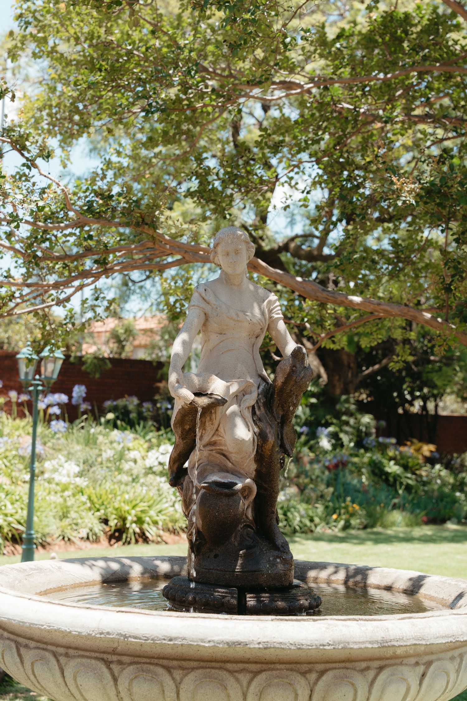 A classical stone fountain with a decorative statue centerpiece stands in a sunny garden with lush trees in the background.