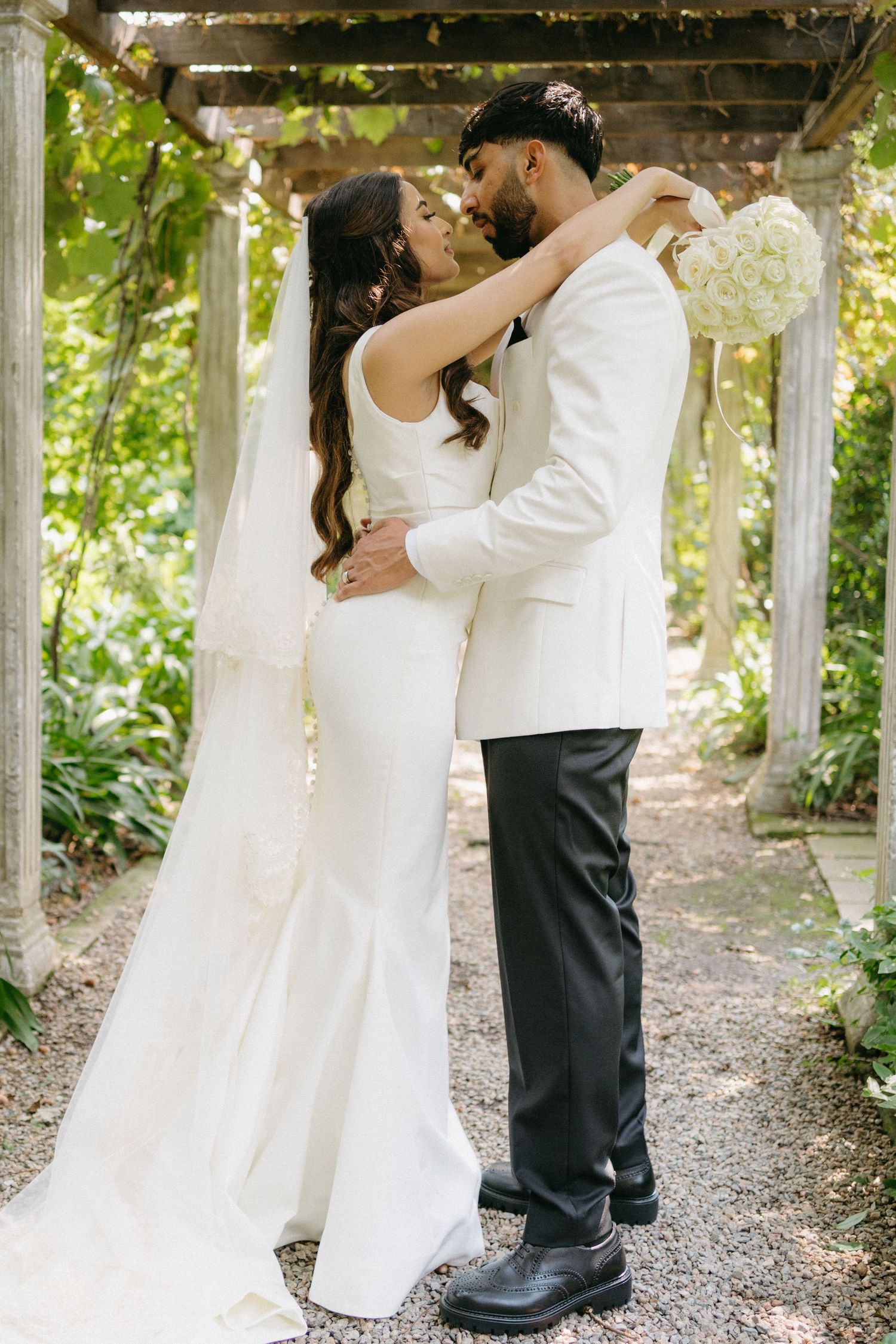 A romantic outdoor wedding moment captured under a pergola with greenery, featuring a couple in elegant white attire and a bridal bouquet.