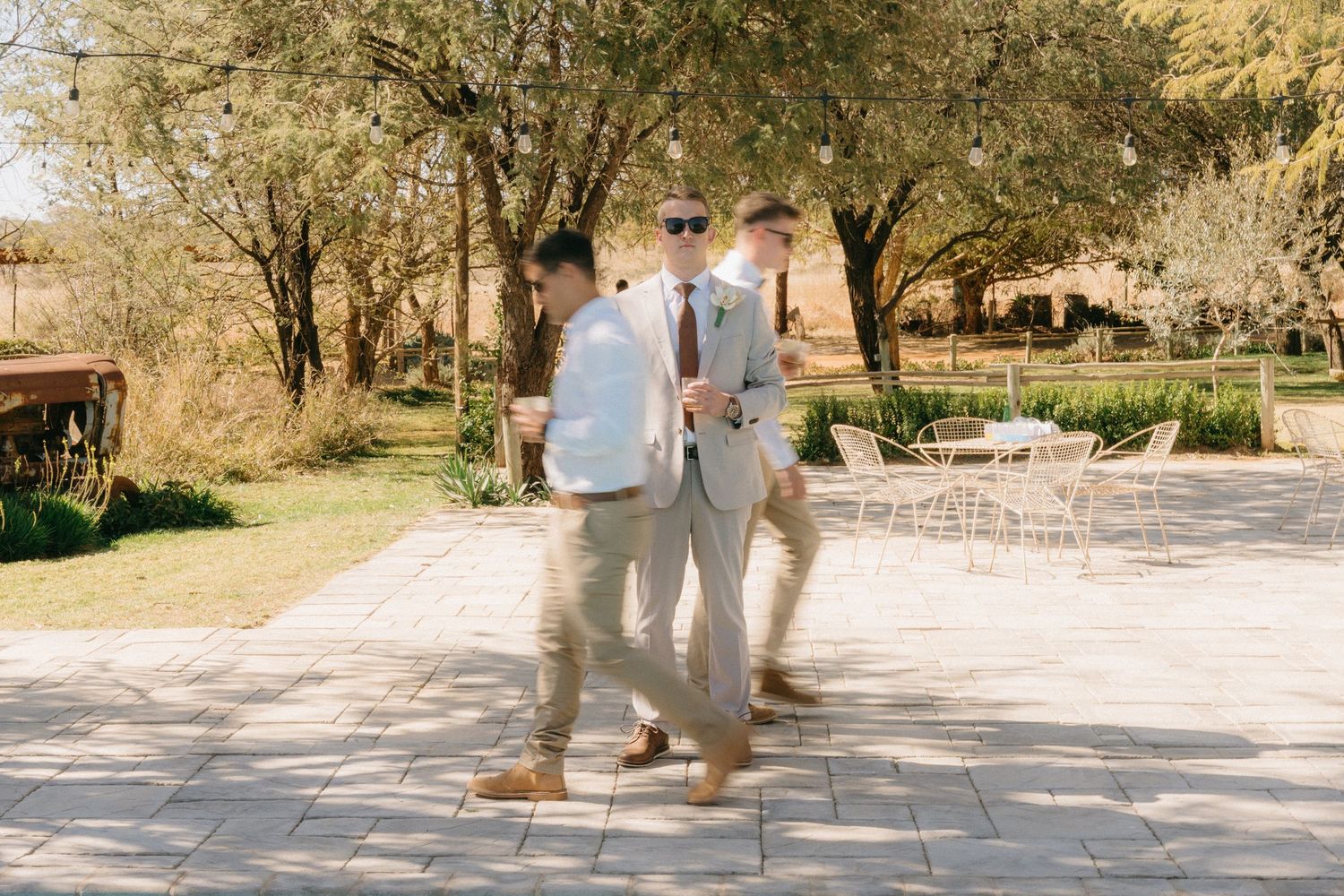 Two people in light colored attire walk together on a stone patio surrounded by trees at an outdoor venue.
