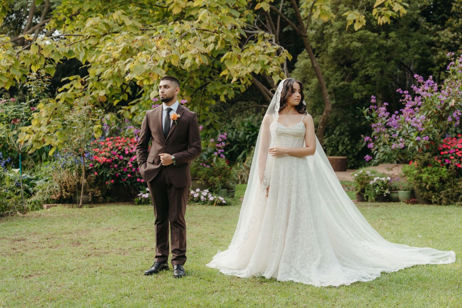 Bride in flowing gown with veil and groom in brown suit standing apart in a lush garden setting in Johannesburg
