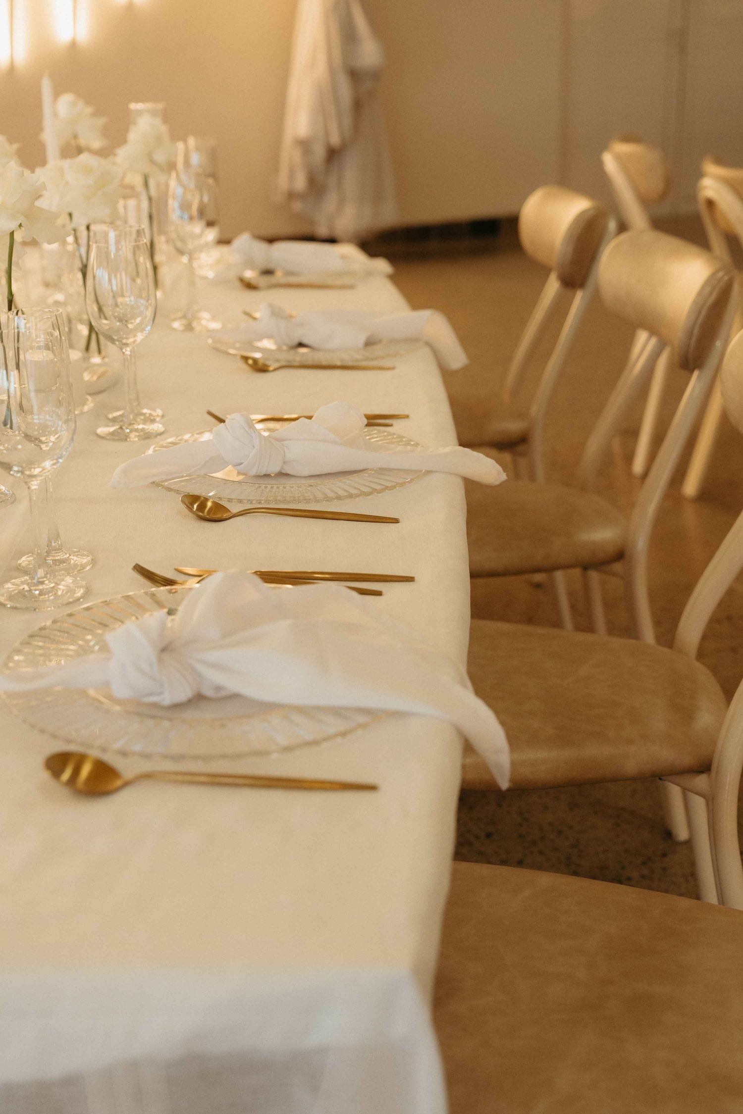 Elegant white wedding reception table setting with gold cutlery, folded napkins, crystal glasses, and white floral centerpieces.