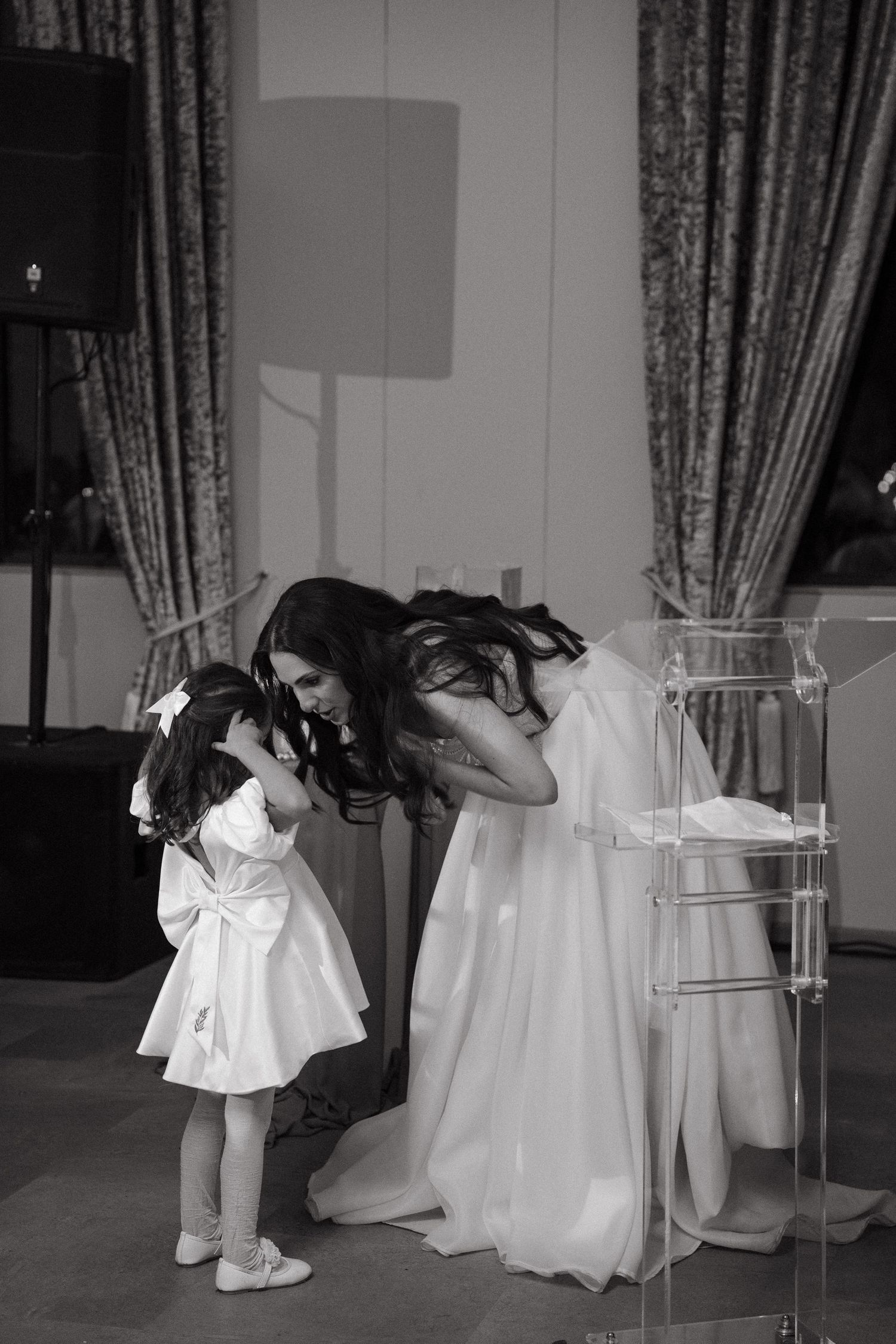 Black and white photo of a bride in a white gown bending down to interact with a young flower girl in a white dress.