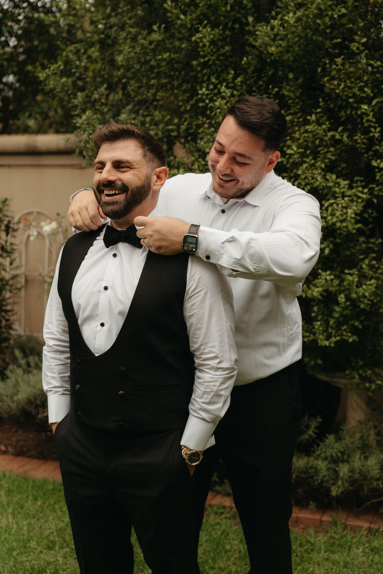 Two men in formal tuxedo attire laugh together outdoors, one adjusting the other's bow tie before a wedding.