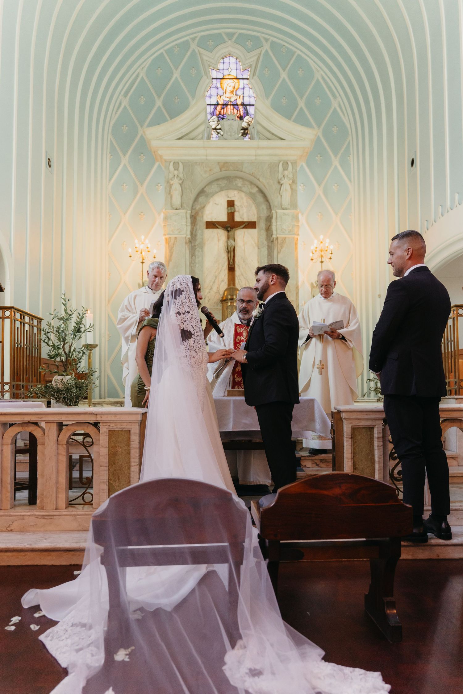 Bride and groom exchange vows at a Catholic church altar, officiated by a priest, with a stunning stained glass window backdrop.