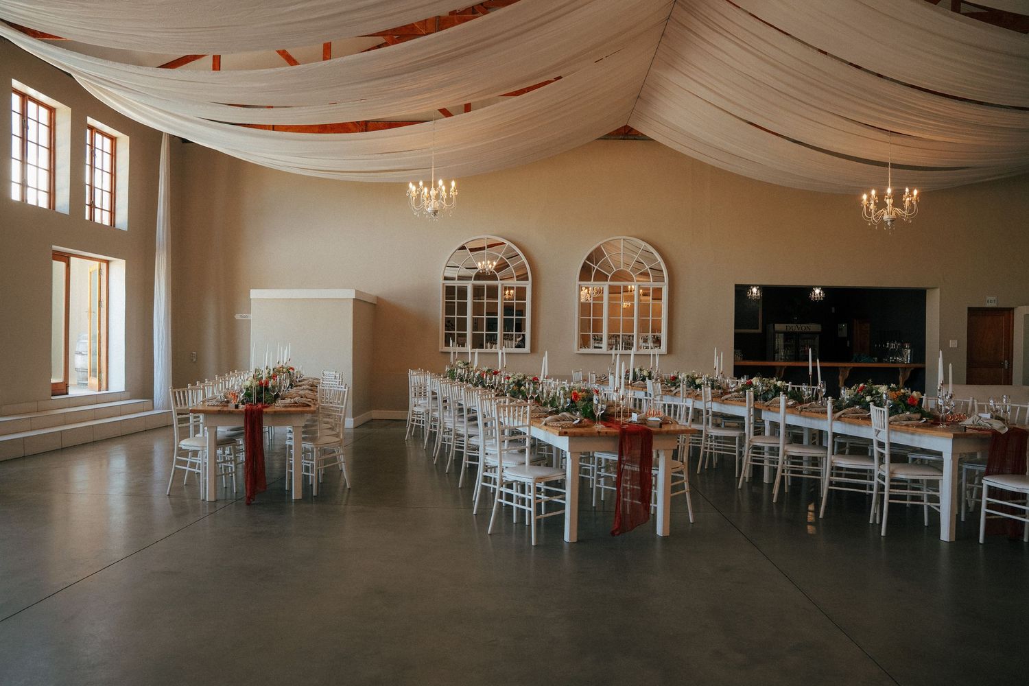 Elegant wedding reception hall with long rustic tables, white chairs, and draped fabric ceiling beneath chandeliers and arched windows.