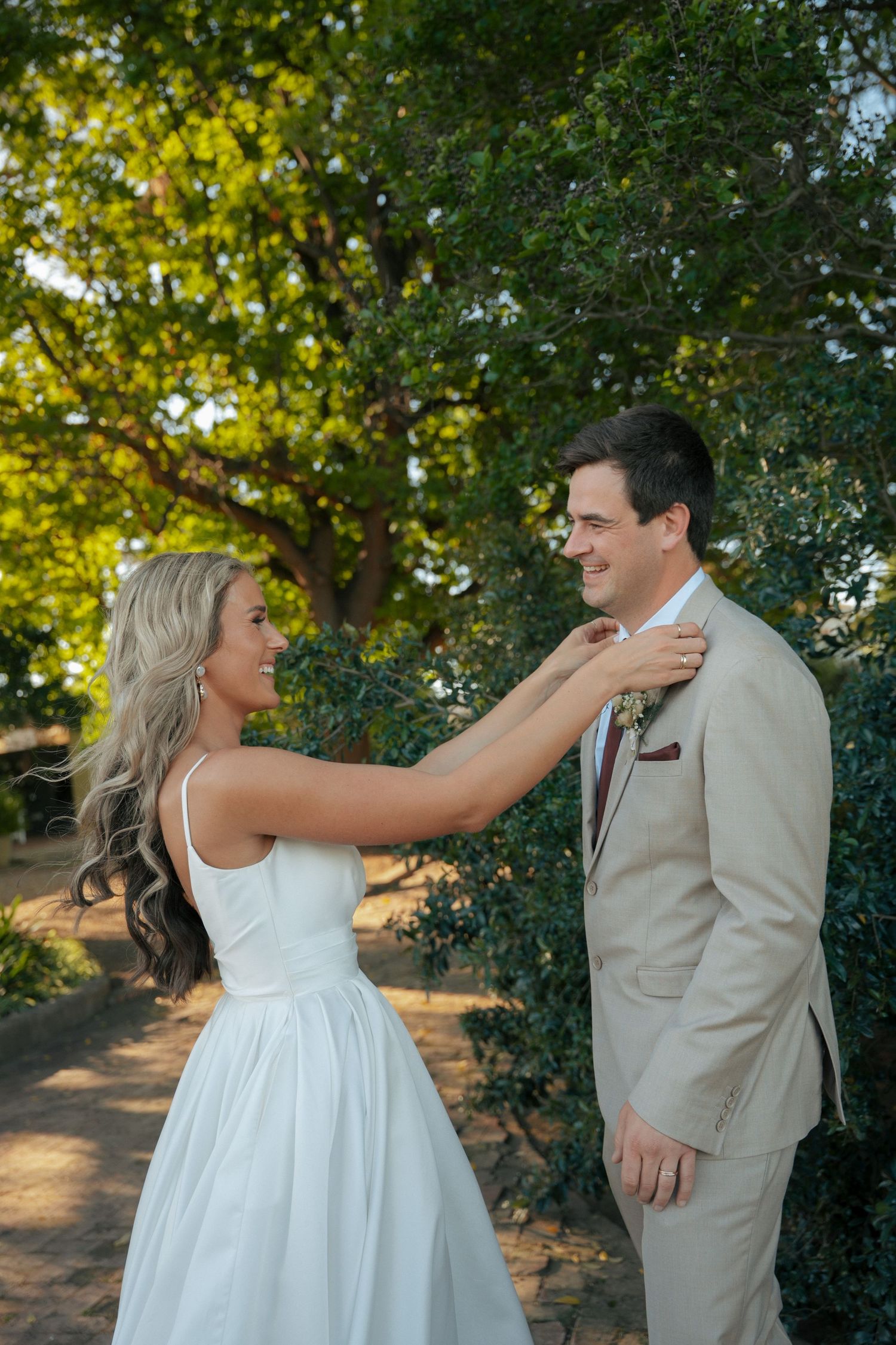 Smiling bride in a white sundress adjusts the boutonniere on her groom's tan suit beneath leafy green trees at an outdoor wedding.