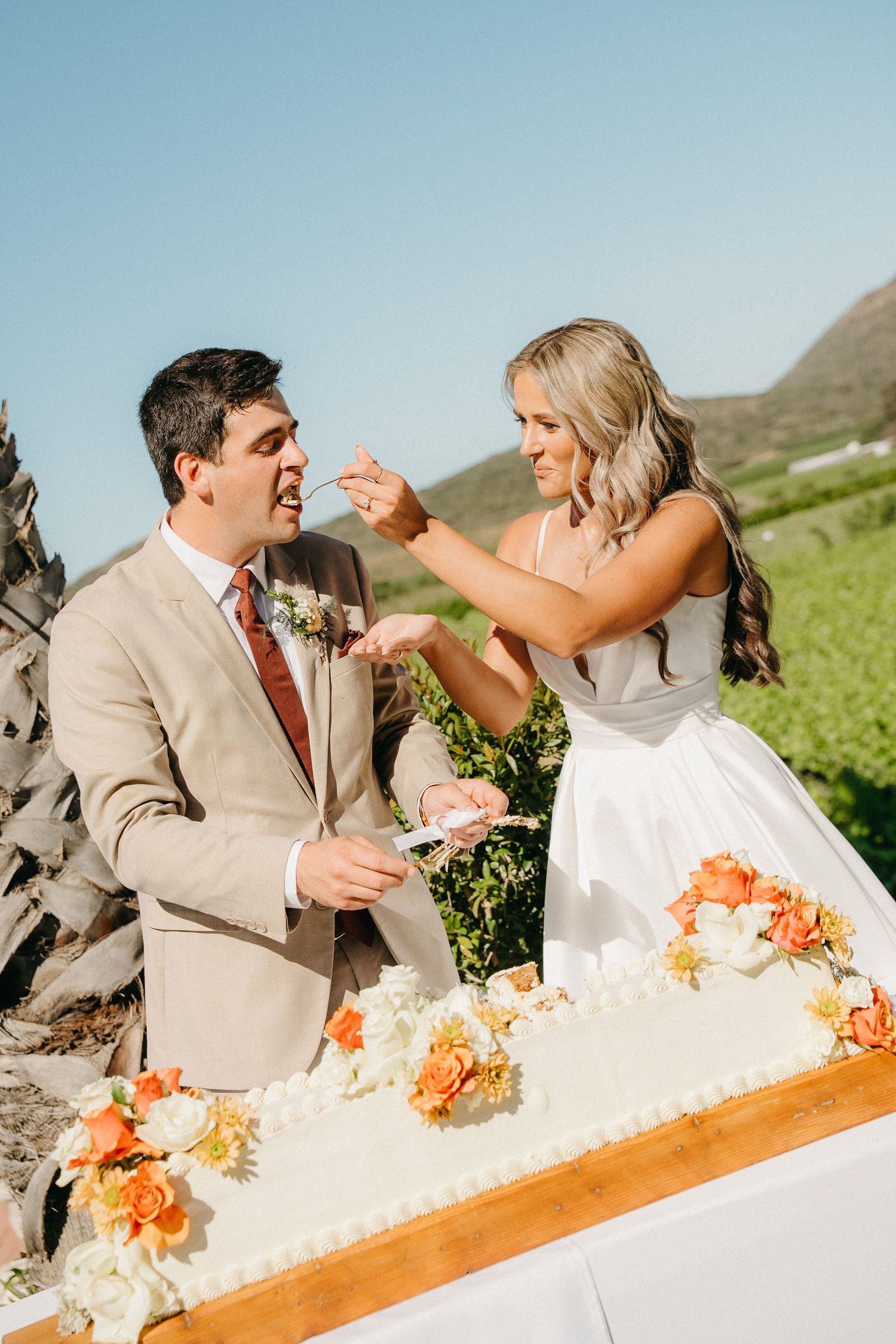Bride in a white gown feeds wedding cake to a smiling groom in a tan suit beside a floral cake with orange and white flowers outdoors.