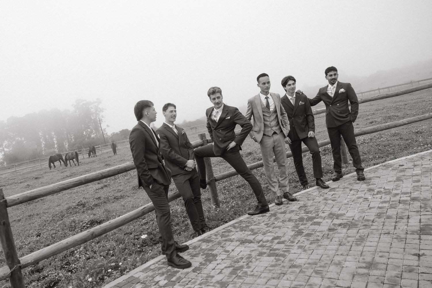 Six young men in suits pose playfully along a wooden fence in a misty outdoor setting, black and white photo.