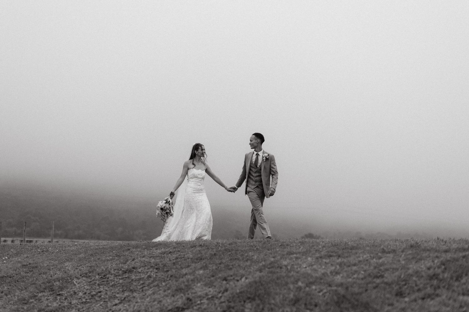 Black and white photo of a bride and groom holding hands walking through misty fog on a hillside.