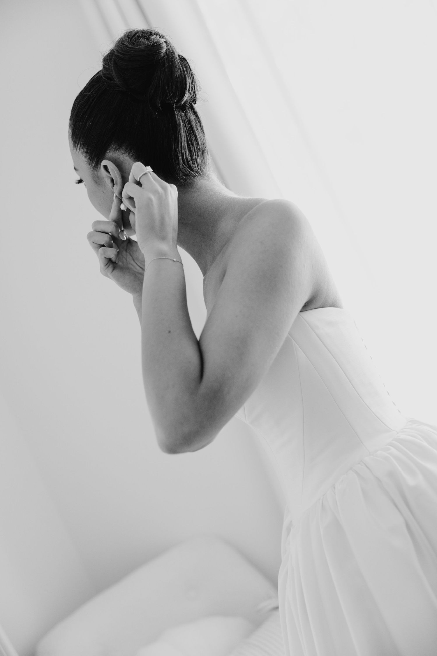 Black and white photo of a bride in a strapless gown putting on earrings while getting ready for her wedding.