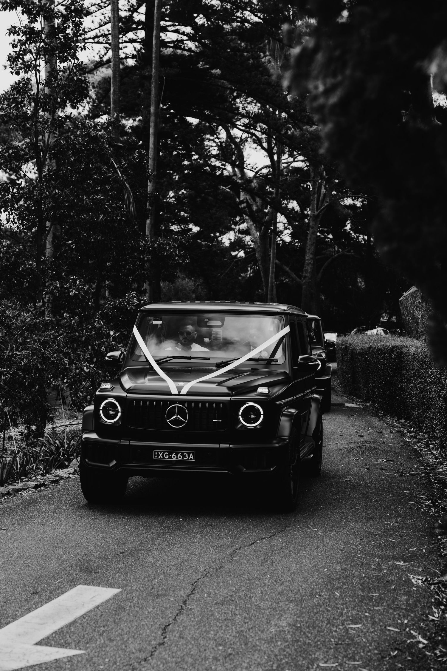 Black Mercedes-Benz G-Class SUV decorated with white wedding ribbons driving down a tree-lined road.