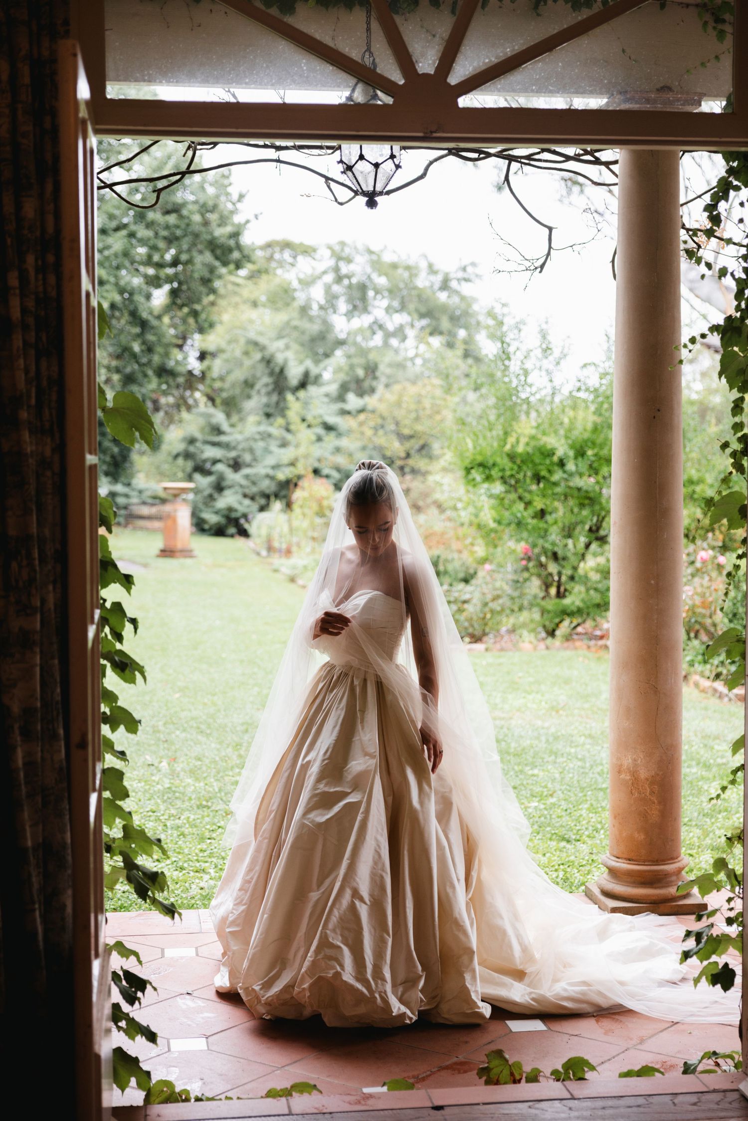 Bride in strapless satin ball gown with long veil stands in elegant vine-covered doorway overlooking garden.