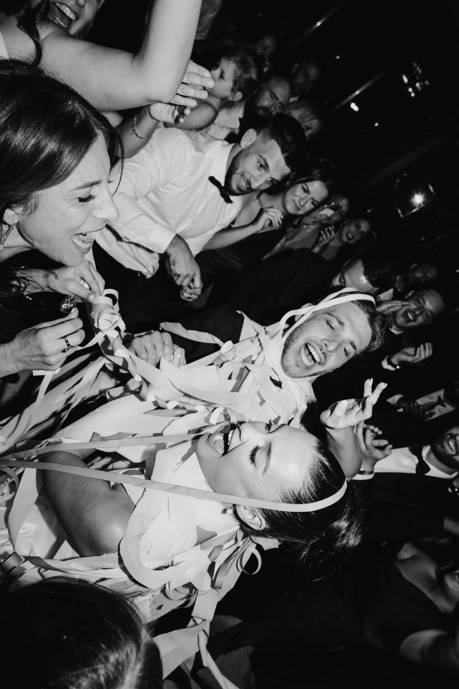 Black and white photo of joyful wedding guests celebrating on the dance floor, wrapped in ribbons.