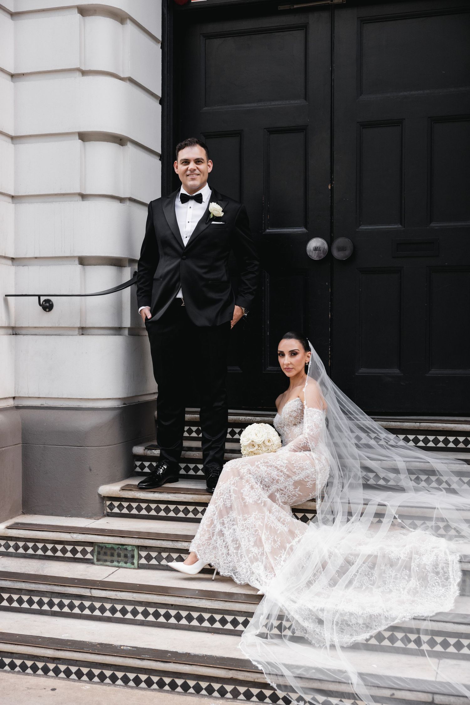 Bride in lace gown sitting on steps with groom standing beside ornate black double doors.