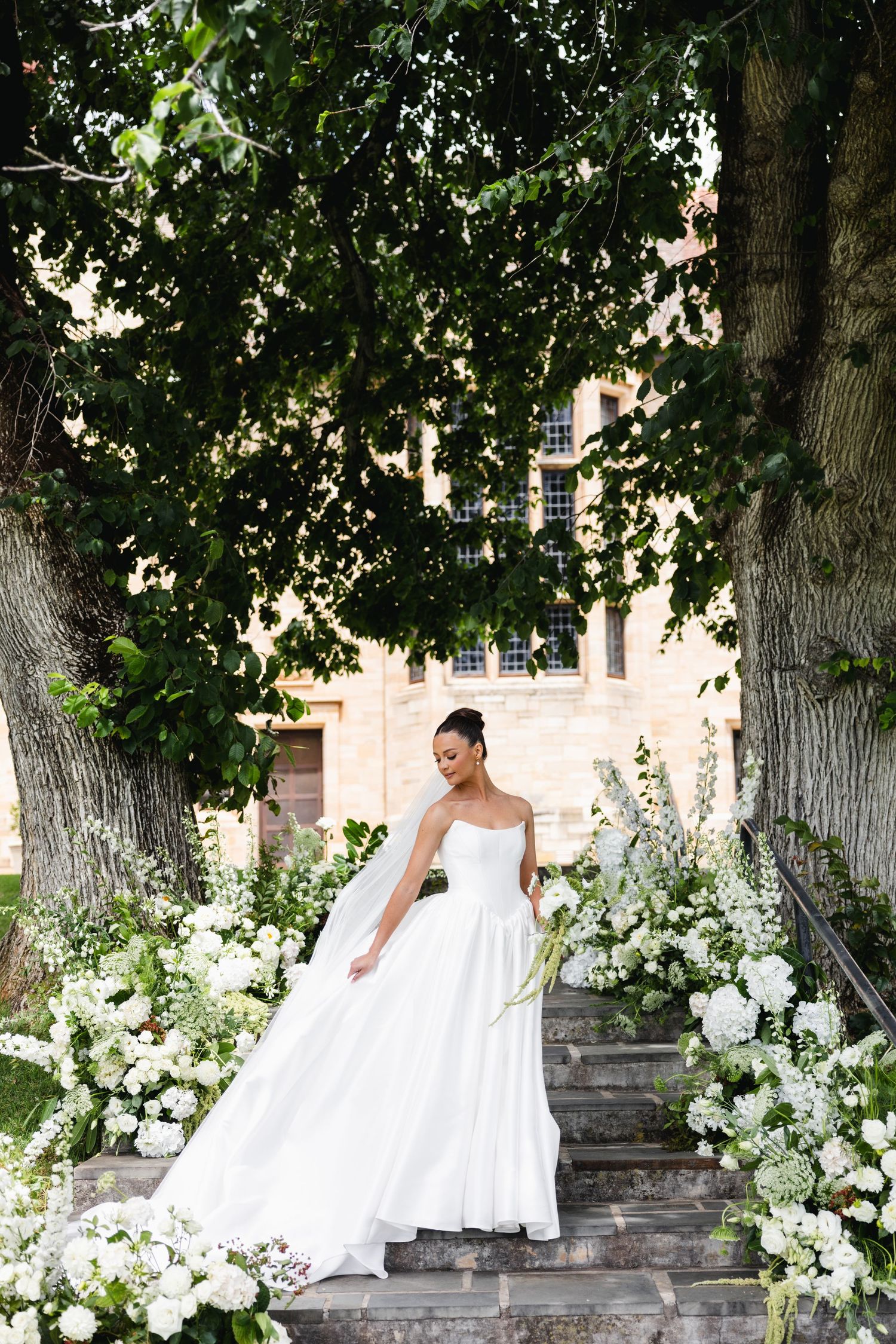 Bride in white strapless ball gown descends flower-lined stone steps beneath lush trees at elegant venue.