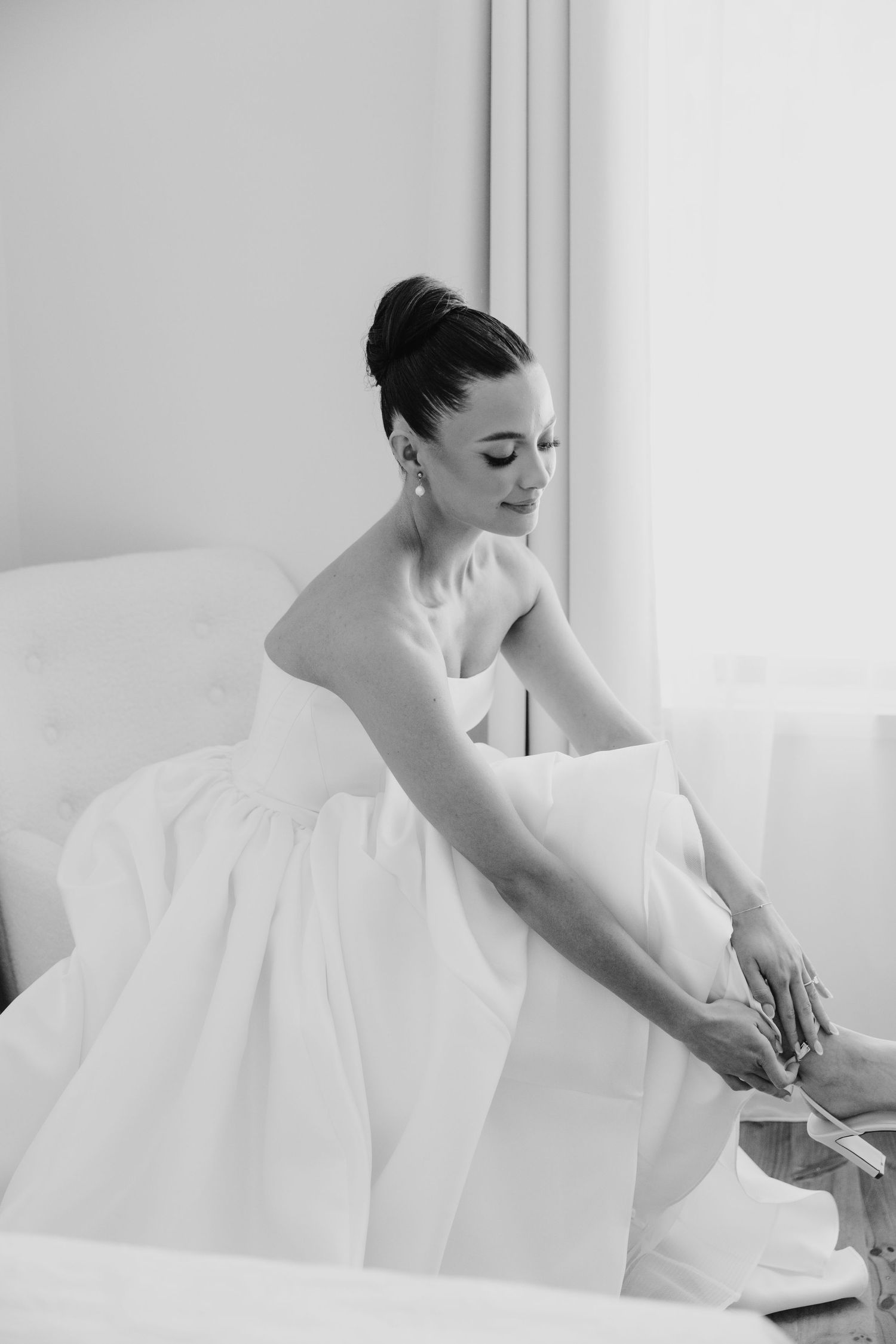 Elegant bride in strapless white ball gown putting on heels while seated near a bright window on wedding day.