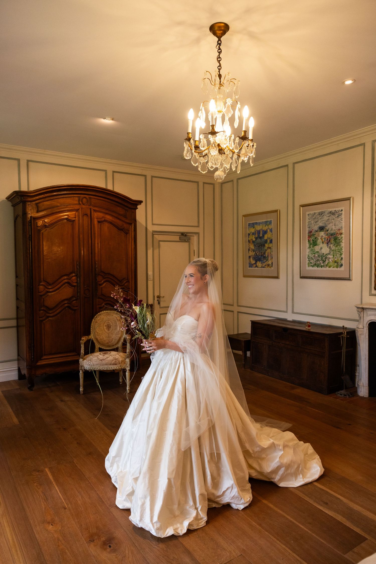 Bride in elegant ivory ballgown with veil holds bouquet in ornate room with crystal chandelier and antique armoire.