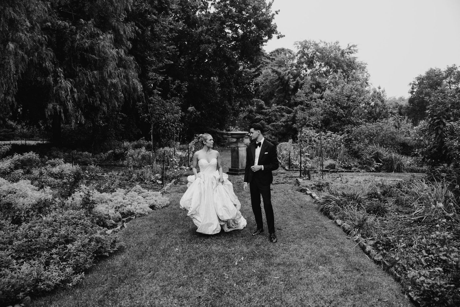 Black and white photo of a bride and groom walking hand in hand through a lush garden on their wedding day.