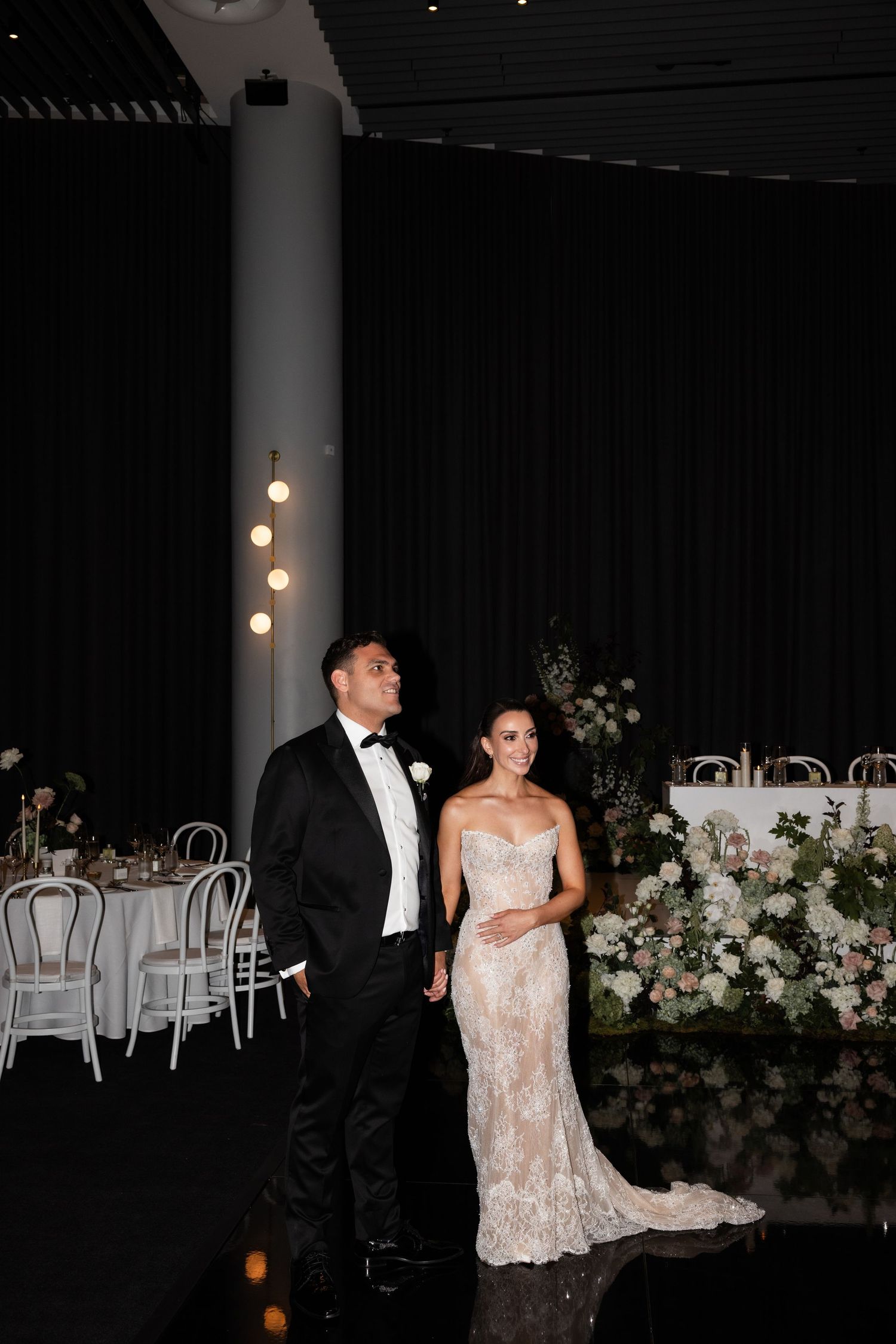 Bride and groom in black tuxedo stand together in elegant dark reception venue with floral arrangements.