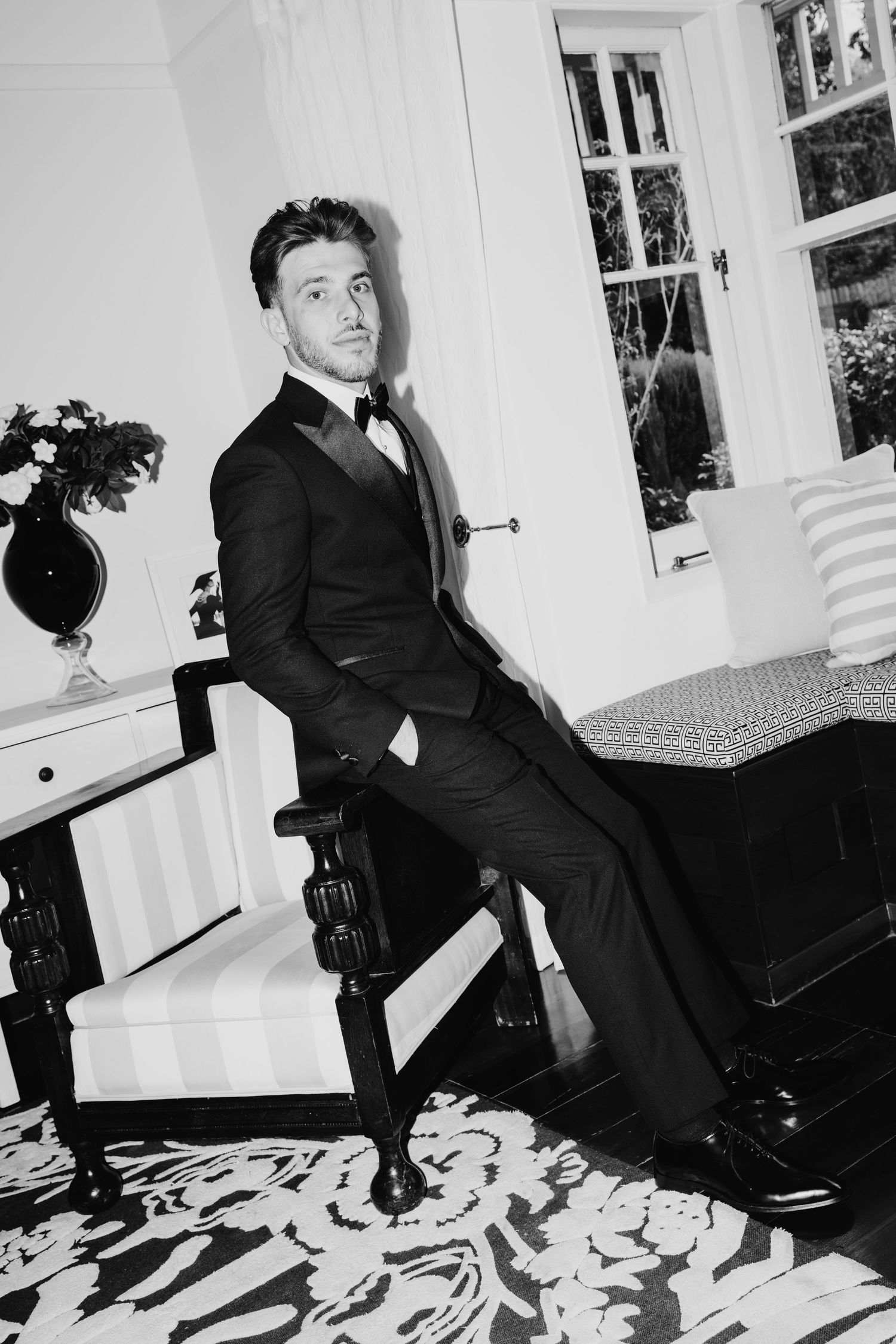A dapper man in a black tuxedo with bow tie sits elegantly in an ornate chair in a sophisticated interior.