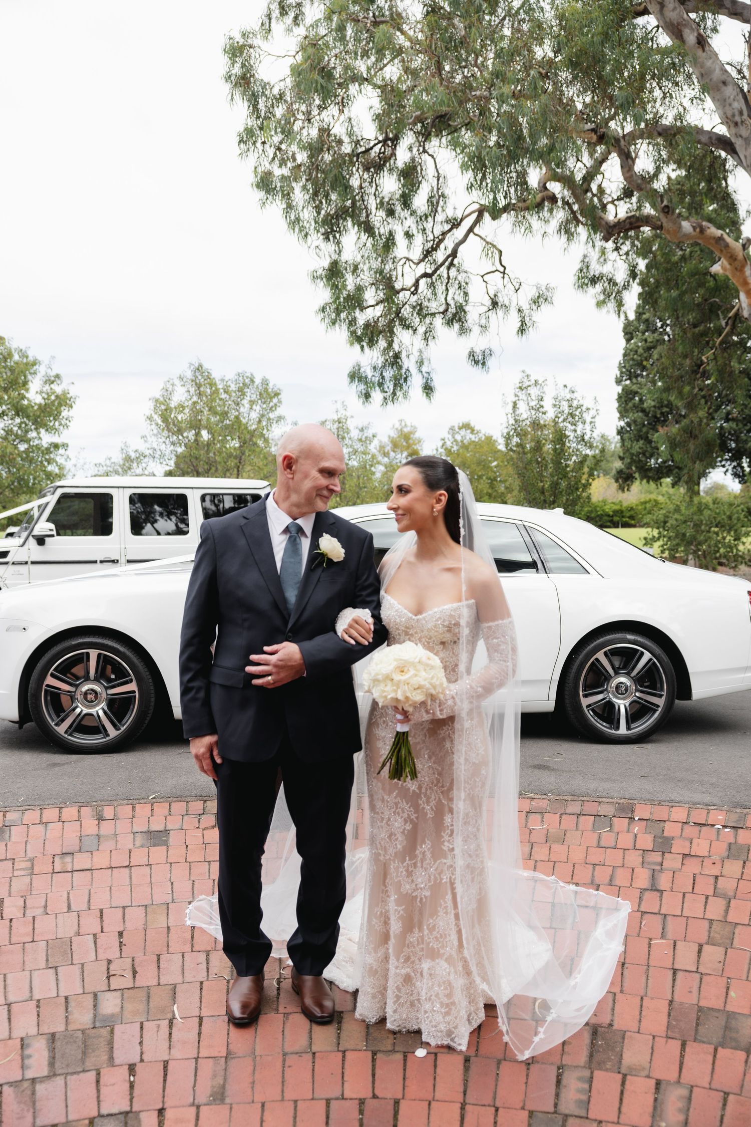 Bride in lace wedding gown with veil stands with older man in suit before white Rolls-Royce on brick plaza.