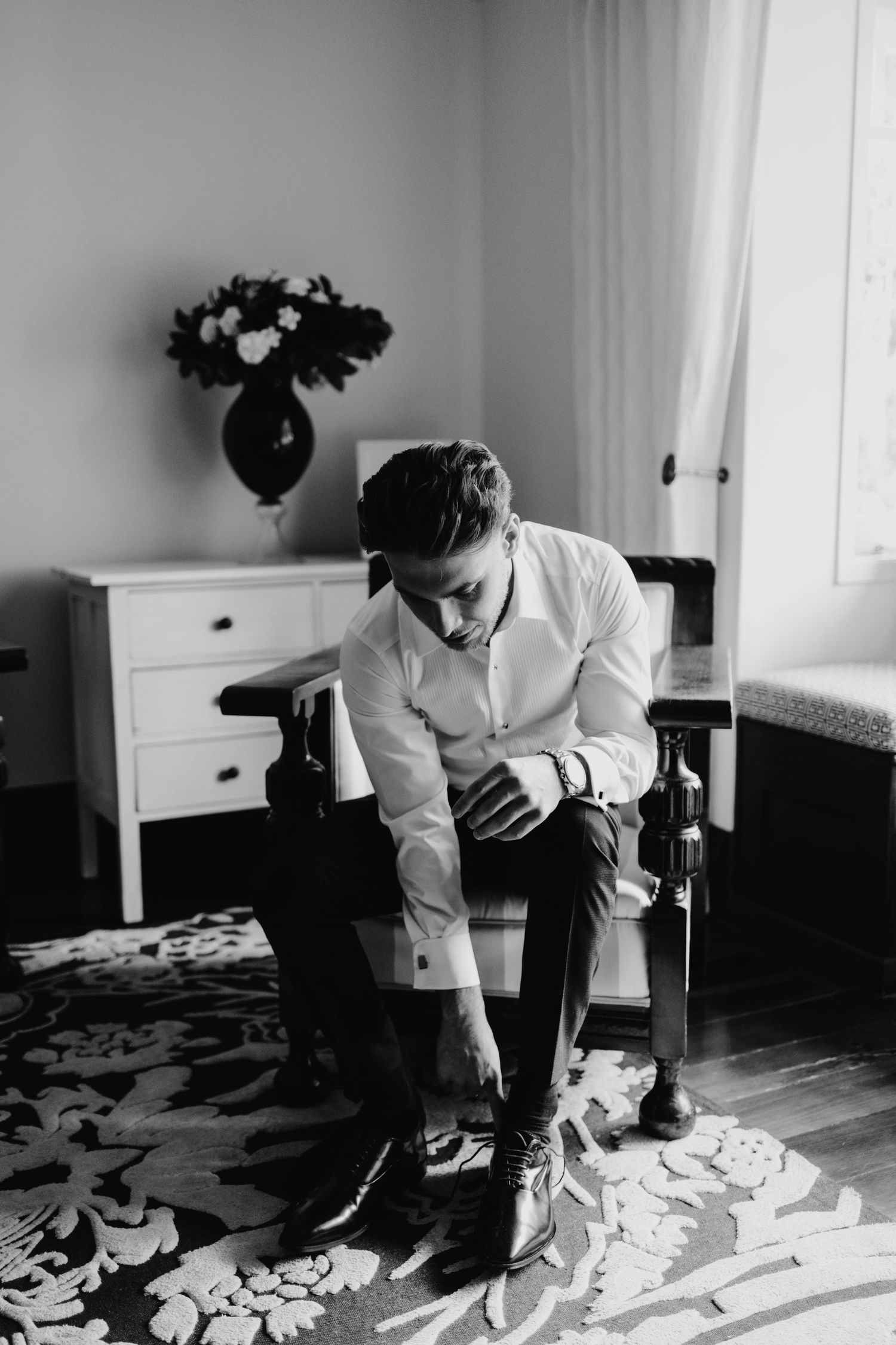 Groom in white dress shirt sitting in chair putting on dress shoes in black and white wedding preparation photo.