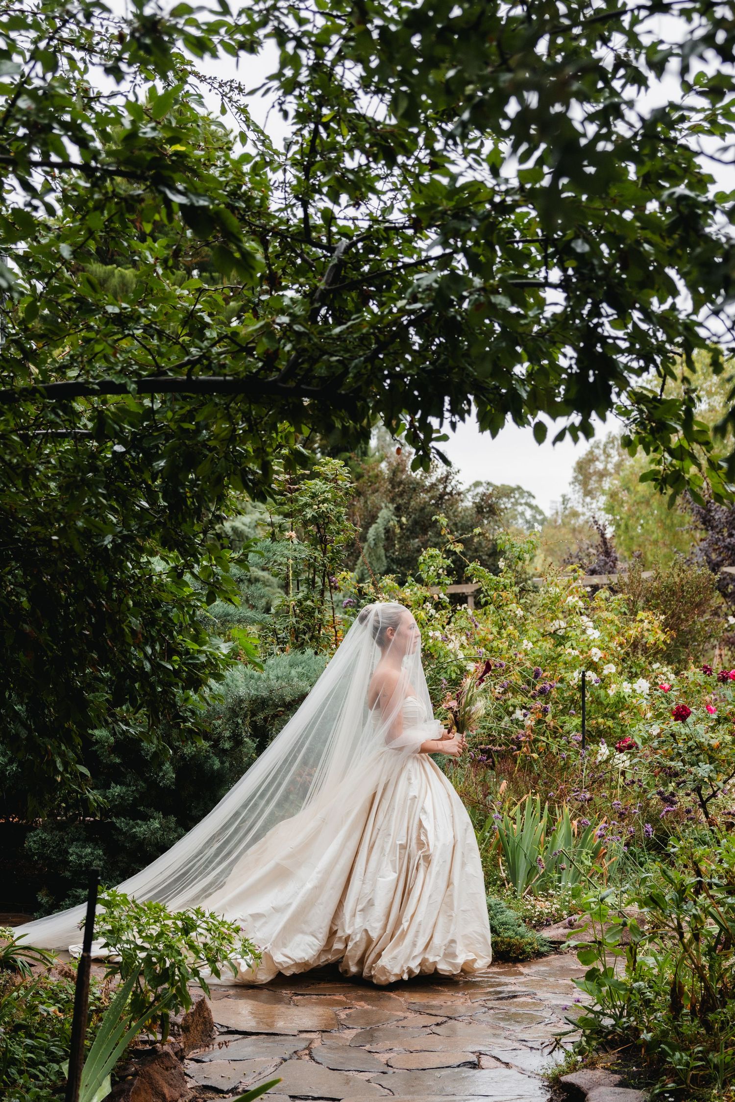 Bride in elegant white gown and flowing veil standing in lush garden surrounded by greenery and blooming flowers.