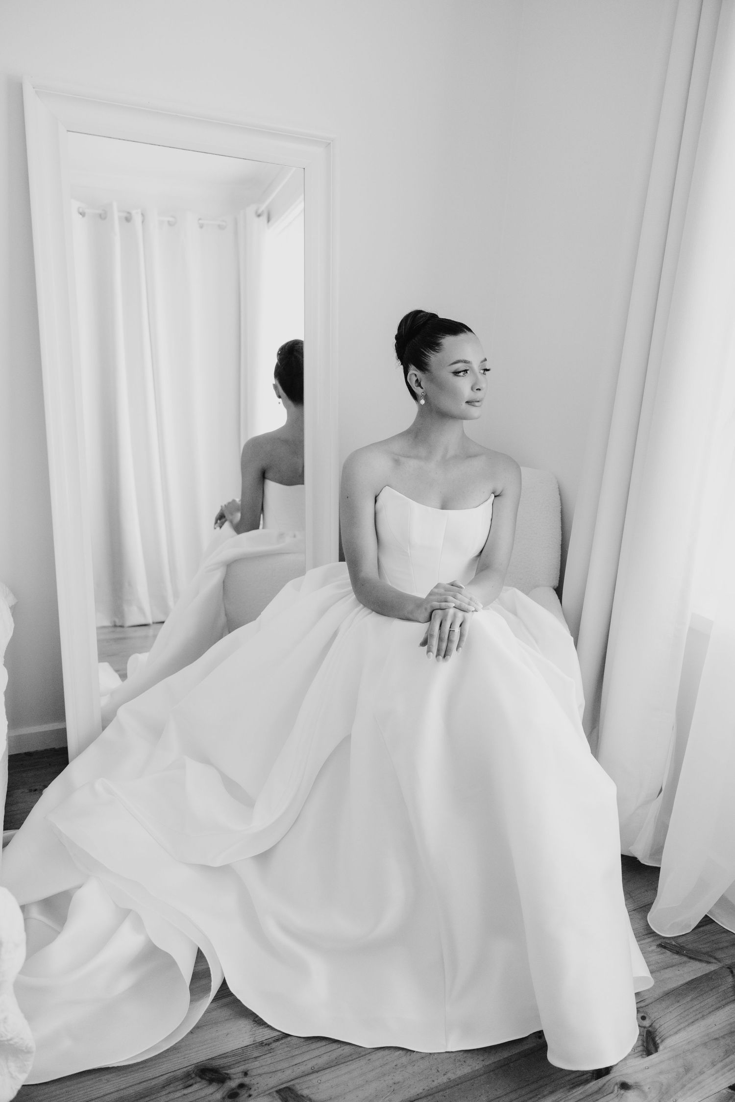 Bride in strapless ball gown seated beside full-length mirror.