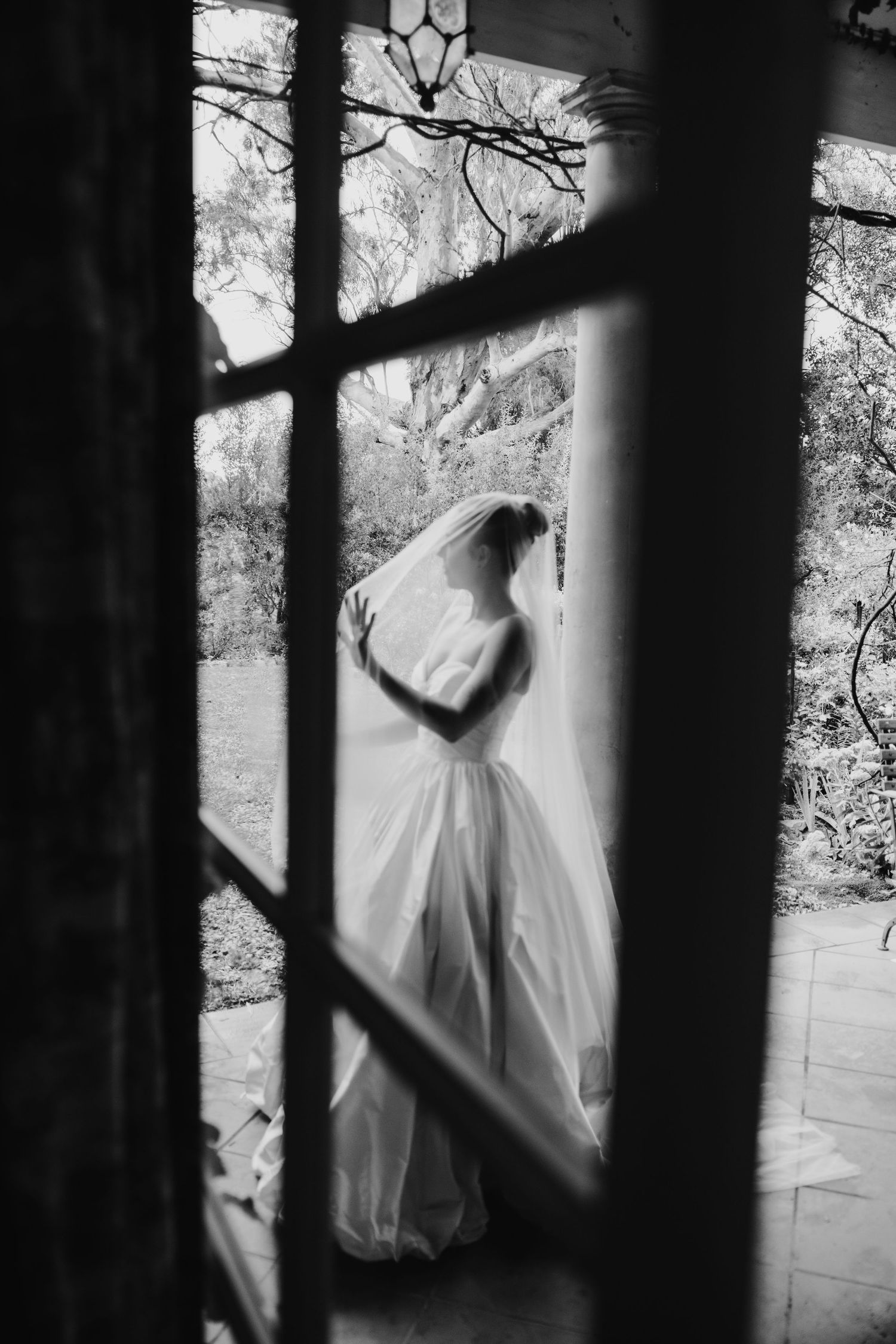 Black and white silhouette of a bride in a wedding gown and veil viewed through a glass-paned door.