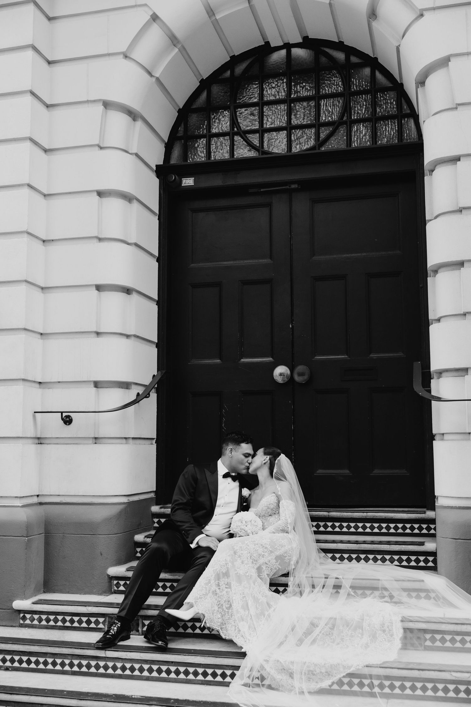 Bride and groom share romantic kiss on church steps, lace veil flowing, in black and white.