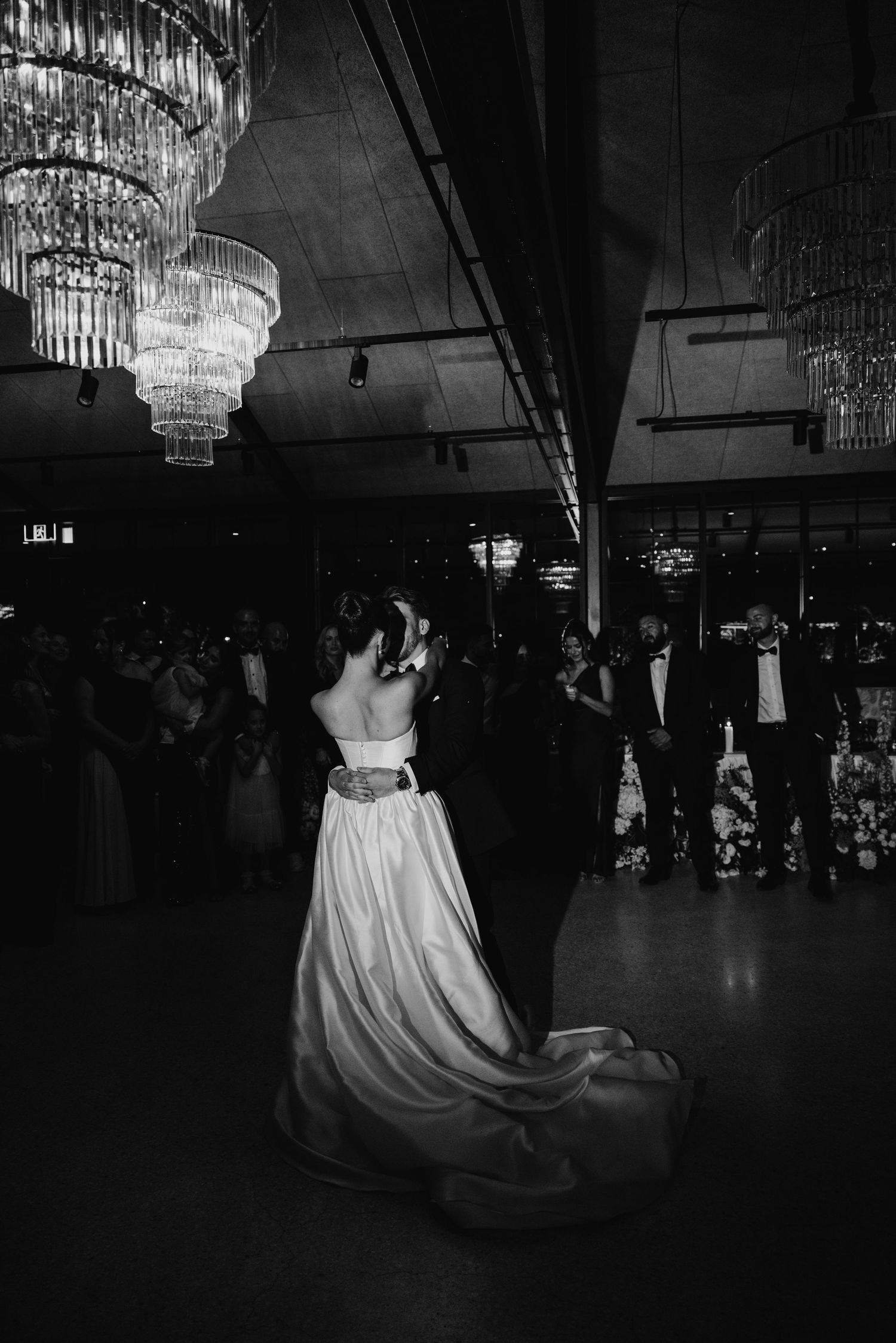 Bride and groom share first dance under crystal chandeliers in elegant black and white wedding reception photo.