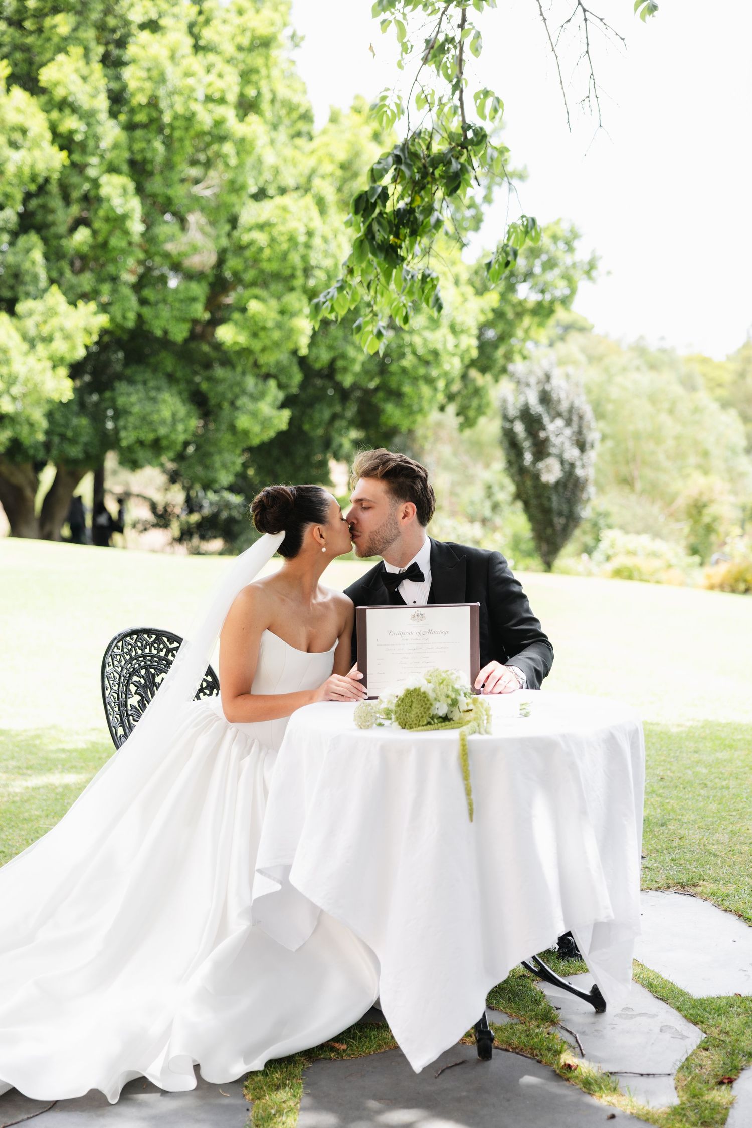 Bride and groom share a kiss while signing wedding registry at elegant outdoor table surrounded by lush greenery.