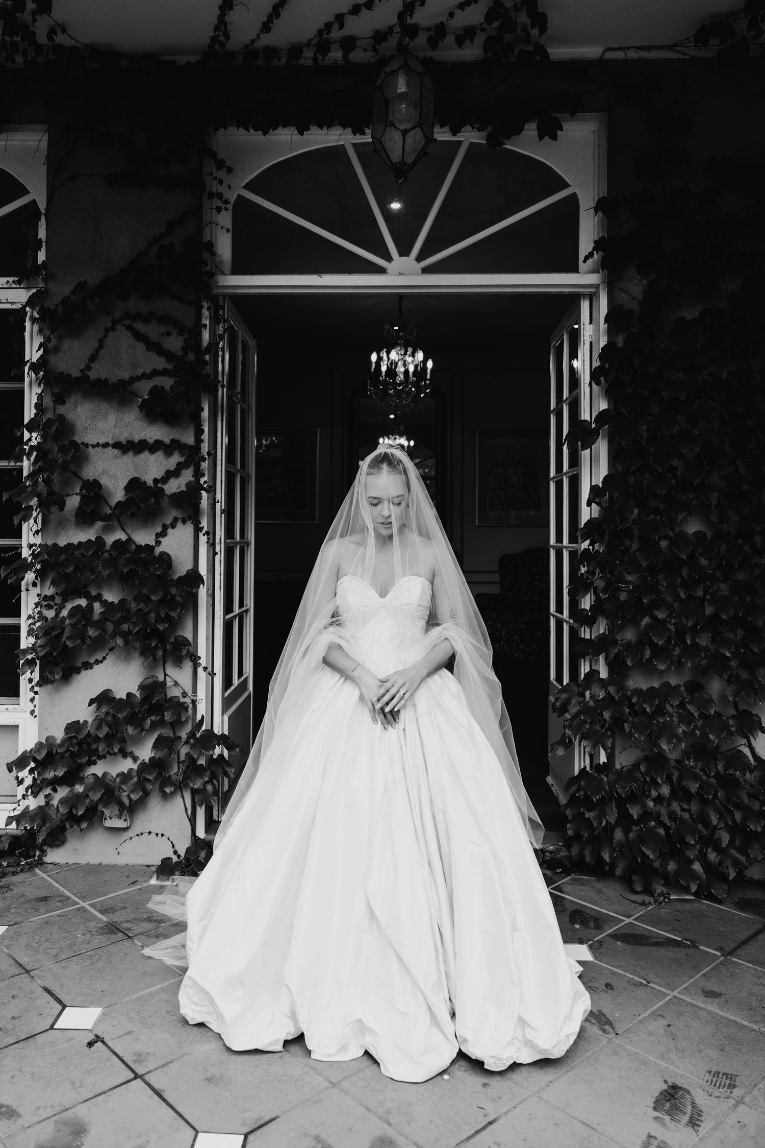 Bride in a strapless ball gown and long veil stands in an ivy-covered doorway with chandelier, black and white photo.