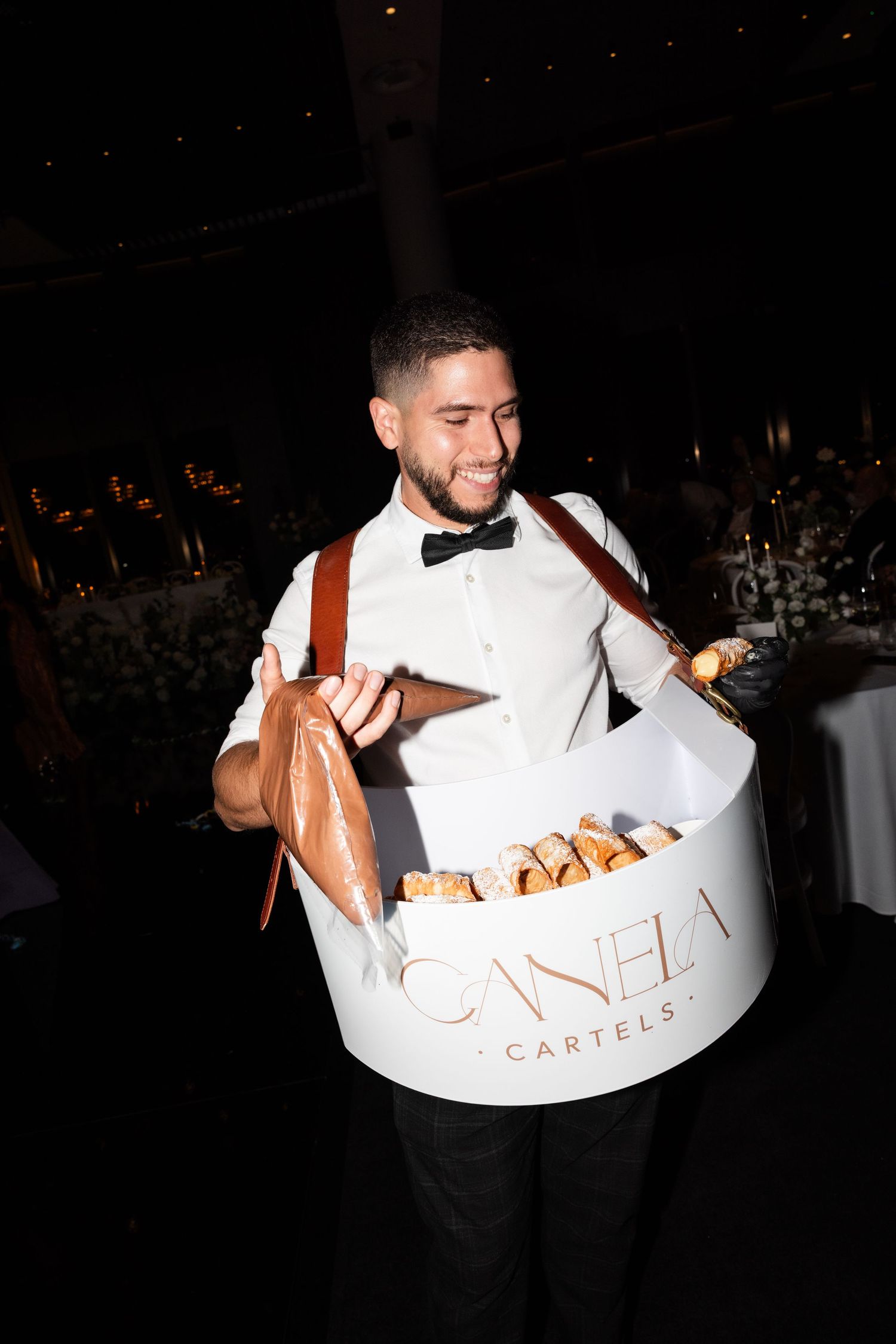 Smiling waiter in white shirt and suspenders carrying a white Canela Cartels bread basket at an elegant event.