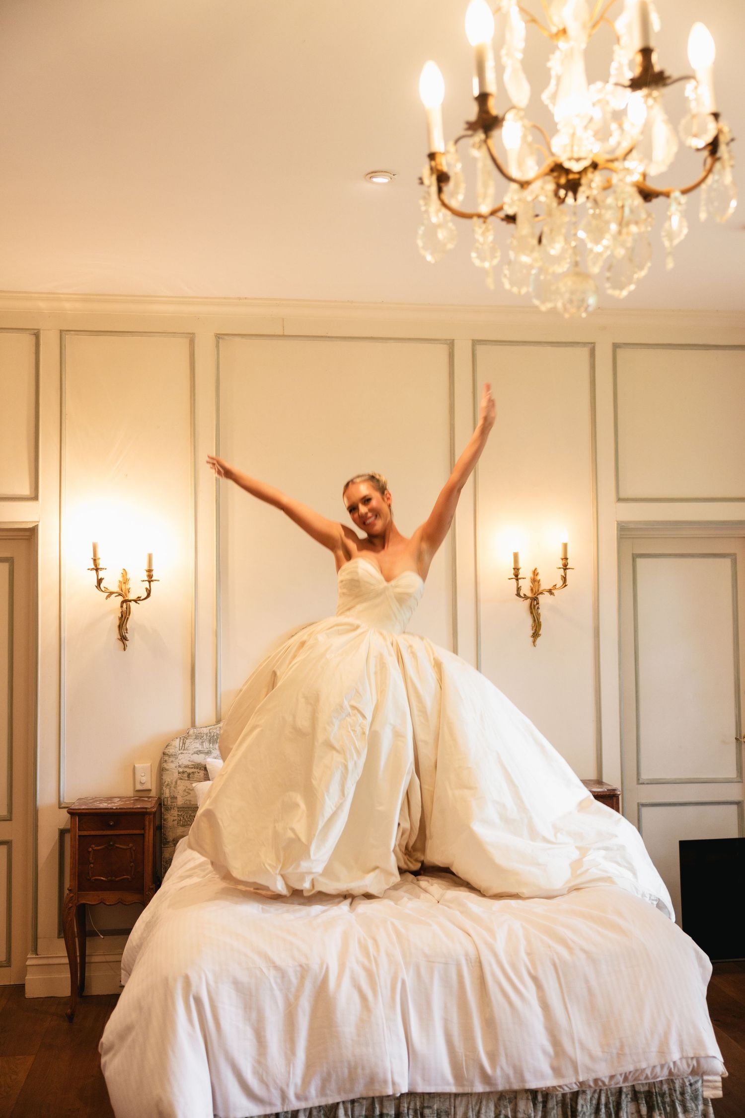 Joyful bride in a voluminous white wedding gown sitting on a bed with arms raised under a crystal chandelier.