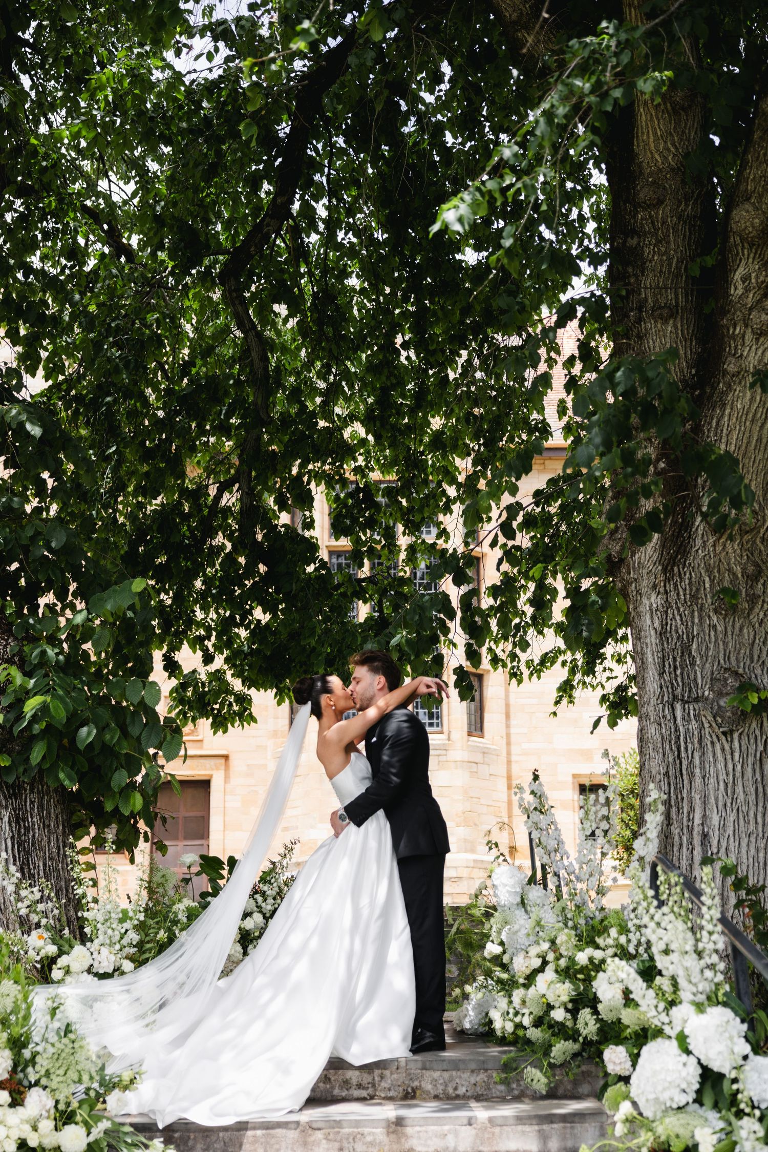 Bride and groom share a romantic kiss under lush green trees, surrounded by white floral arrangements at an outdoor wedding.