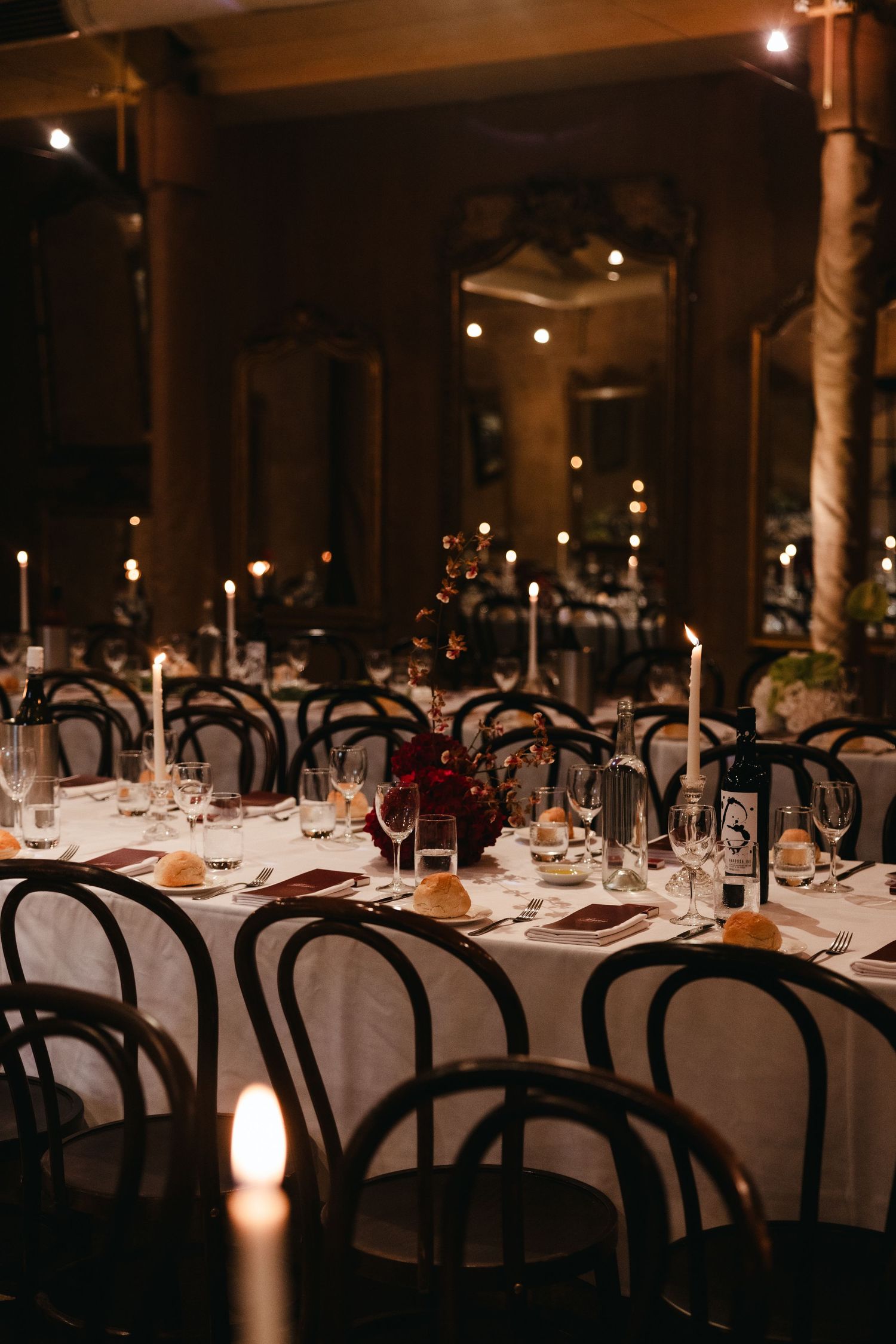 Elegant candlelit wedding reception with black bentwood chairs, white tablecloths, tall tapers, and deep red floral centerpieces.