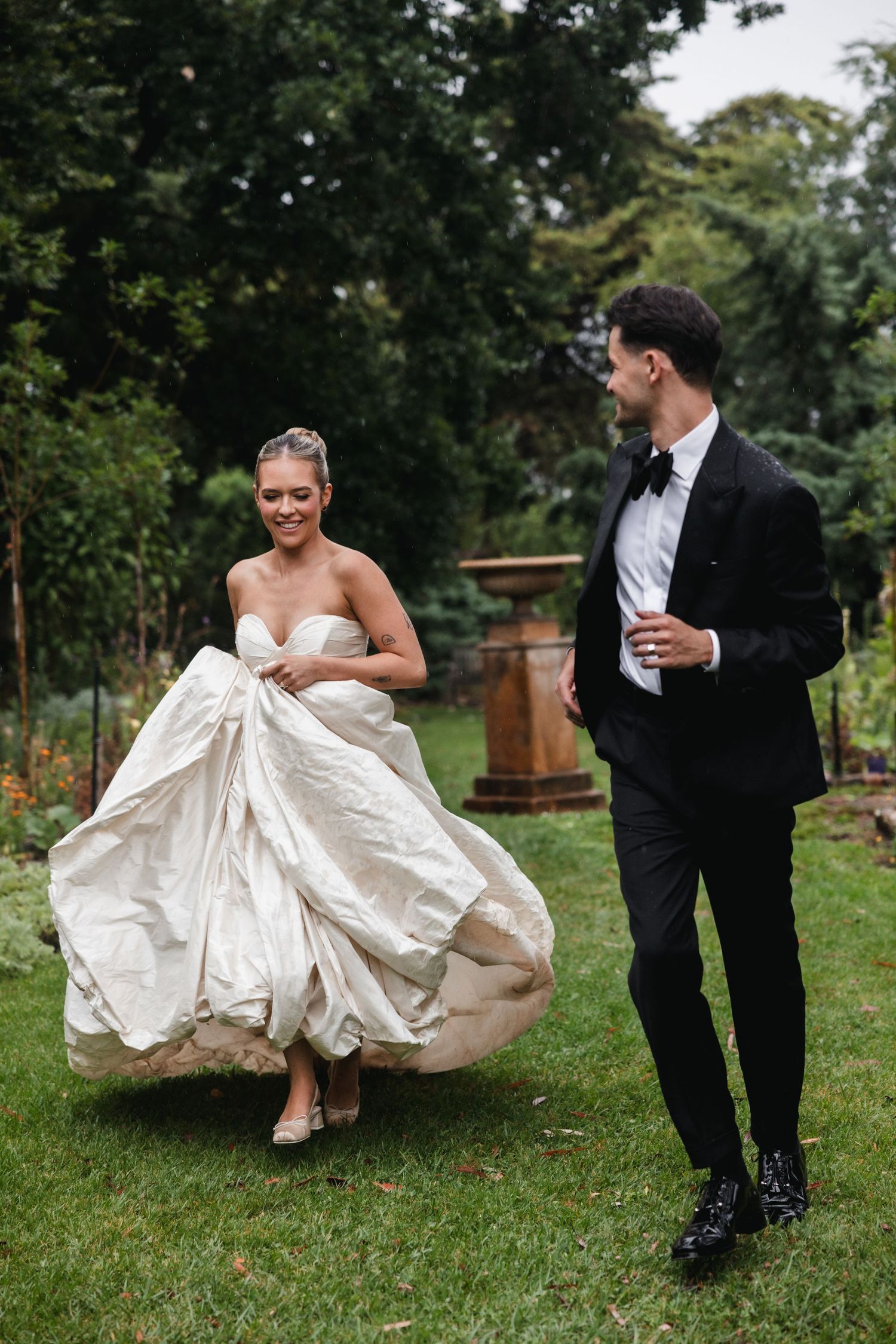 Bride in strapless ball gown lifts dress while walking with groom in black tuxedo through lush garden.
