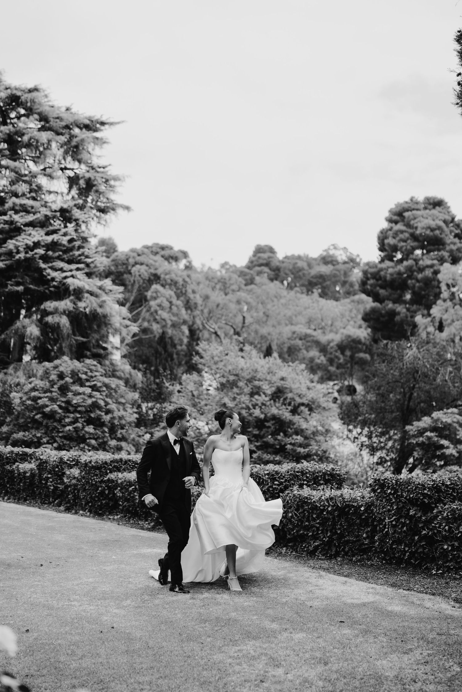 Black and white photo of bride and groom walking hand in hand through lush garden grounds on their wedding day.