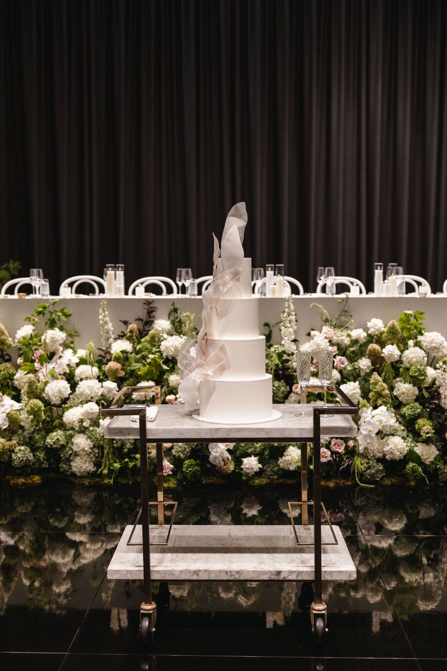 Elegant three-tier white wedding cake with feather topper displayed on a gold cart surrounded by floral arrangements.