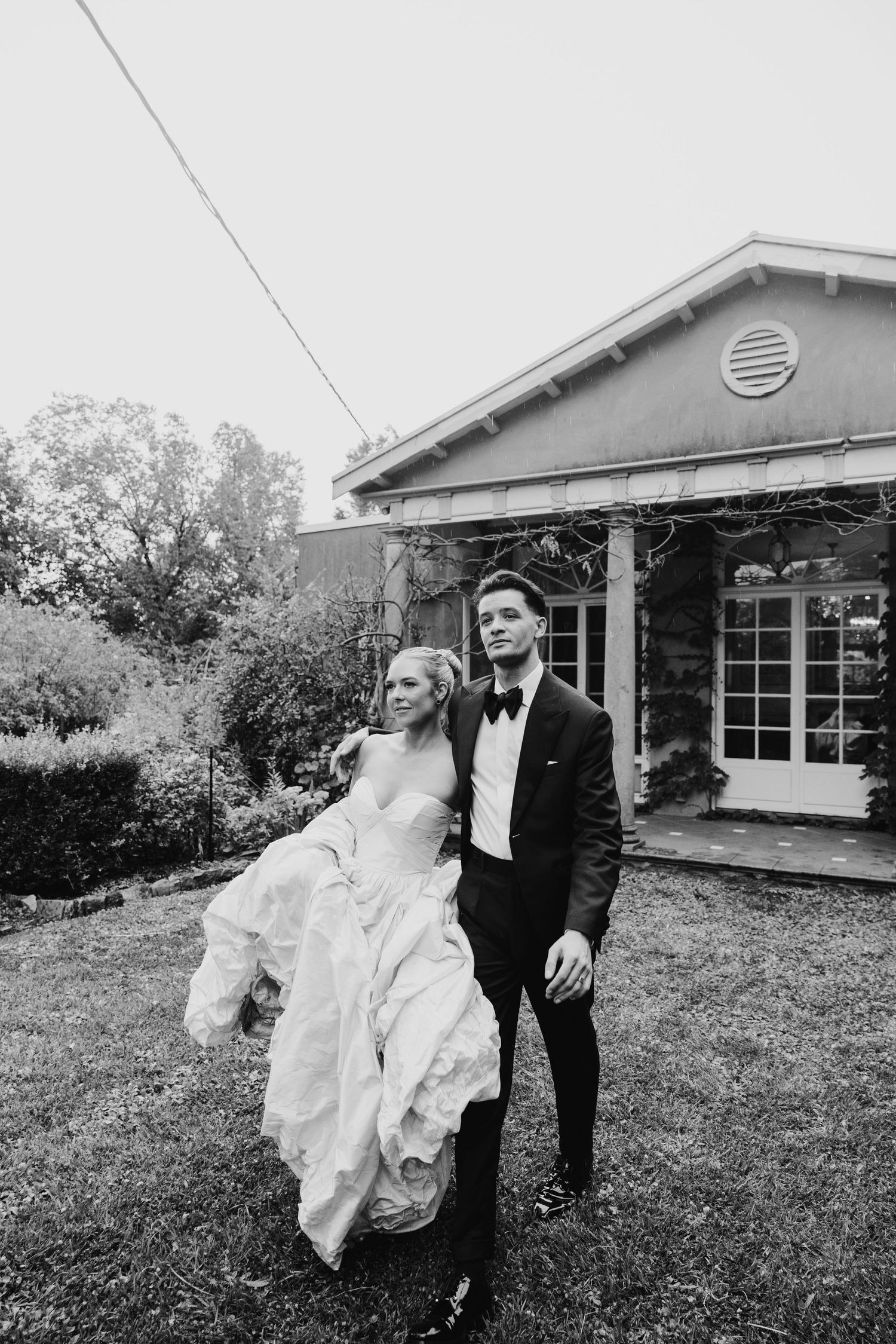 Black and white photo of a bride and groom posing outdoors in front of a house on their wedding day.